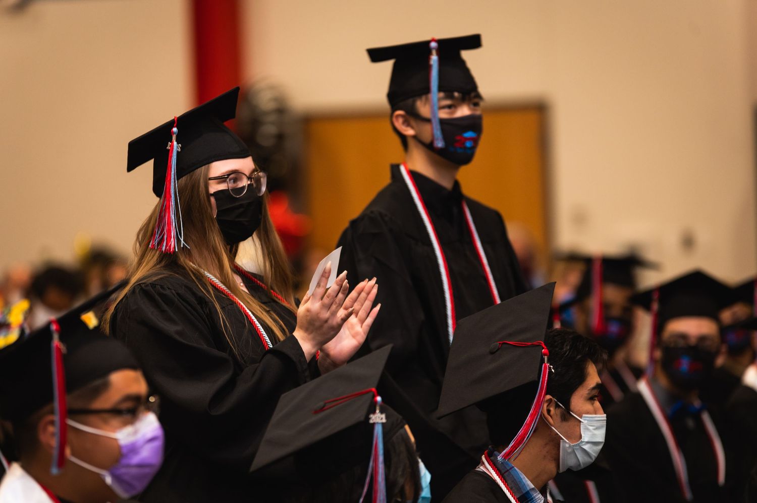 Graduates in black gowns and face masks applauding during ceremony.