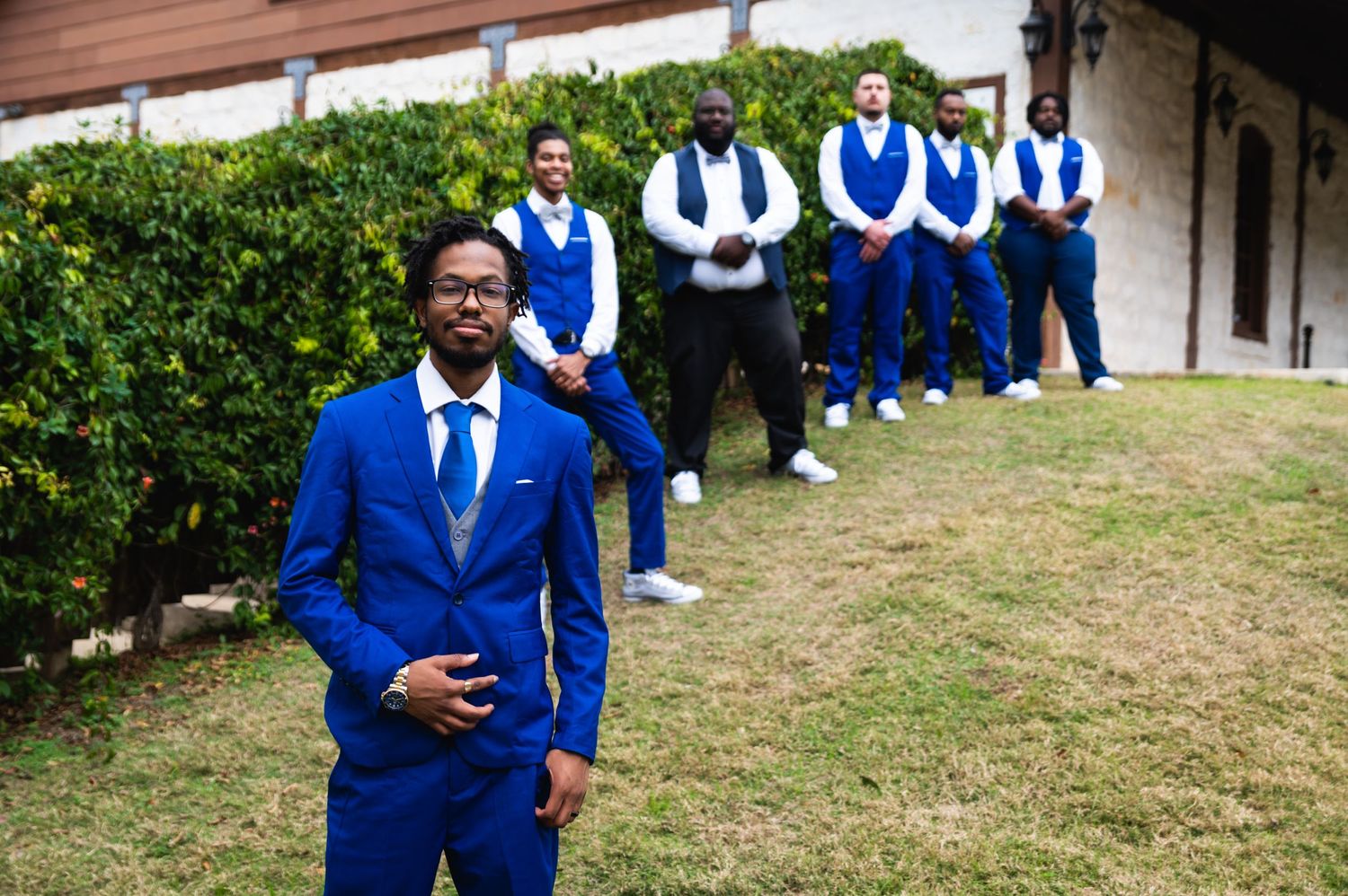 A formal group photo of groomsmen in matching royal blue suits posed against greenery.