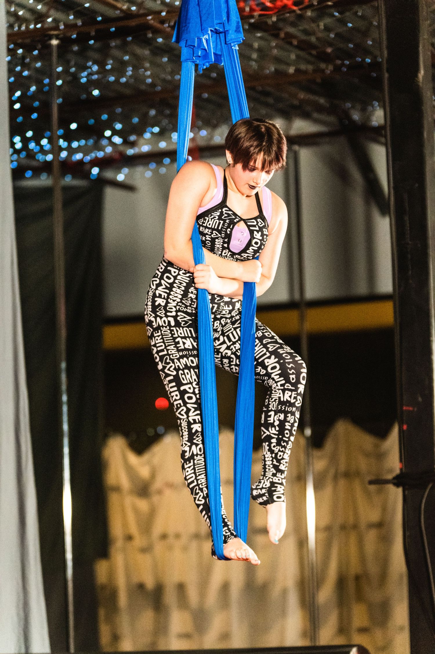 Performer in black and white patterned outfit poses on blue aerial silks during performance.