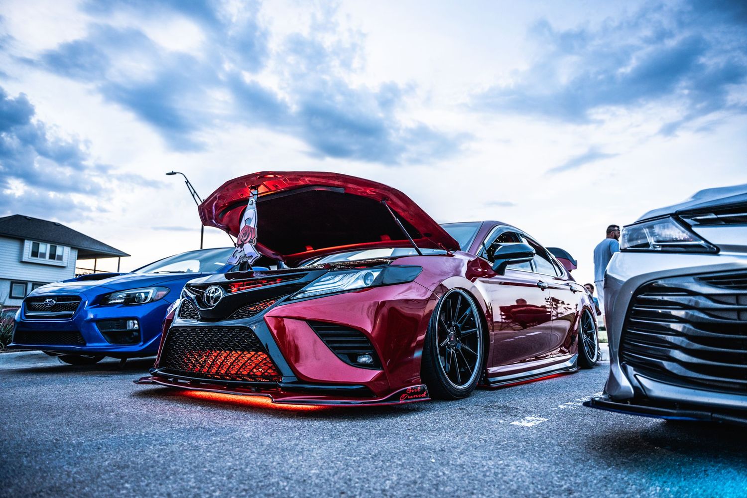 Lowered red Toyota Camry with aggressive body kit flanked by modified cars at dusk.