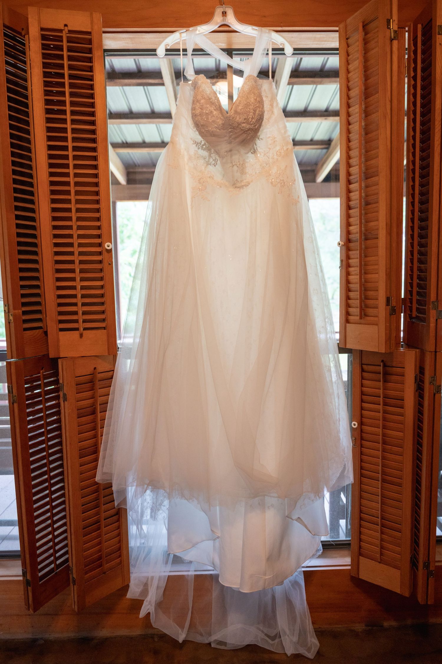 Elegant white wedding dress with lace bodice and tulle skirt hanging in wooden shuttered window.