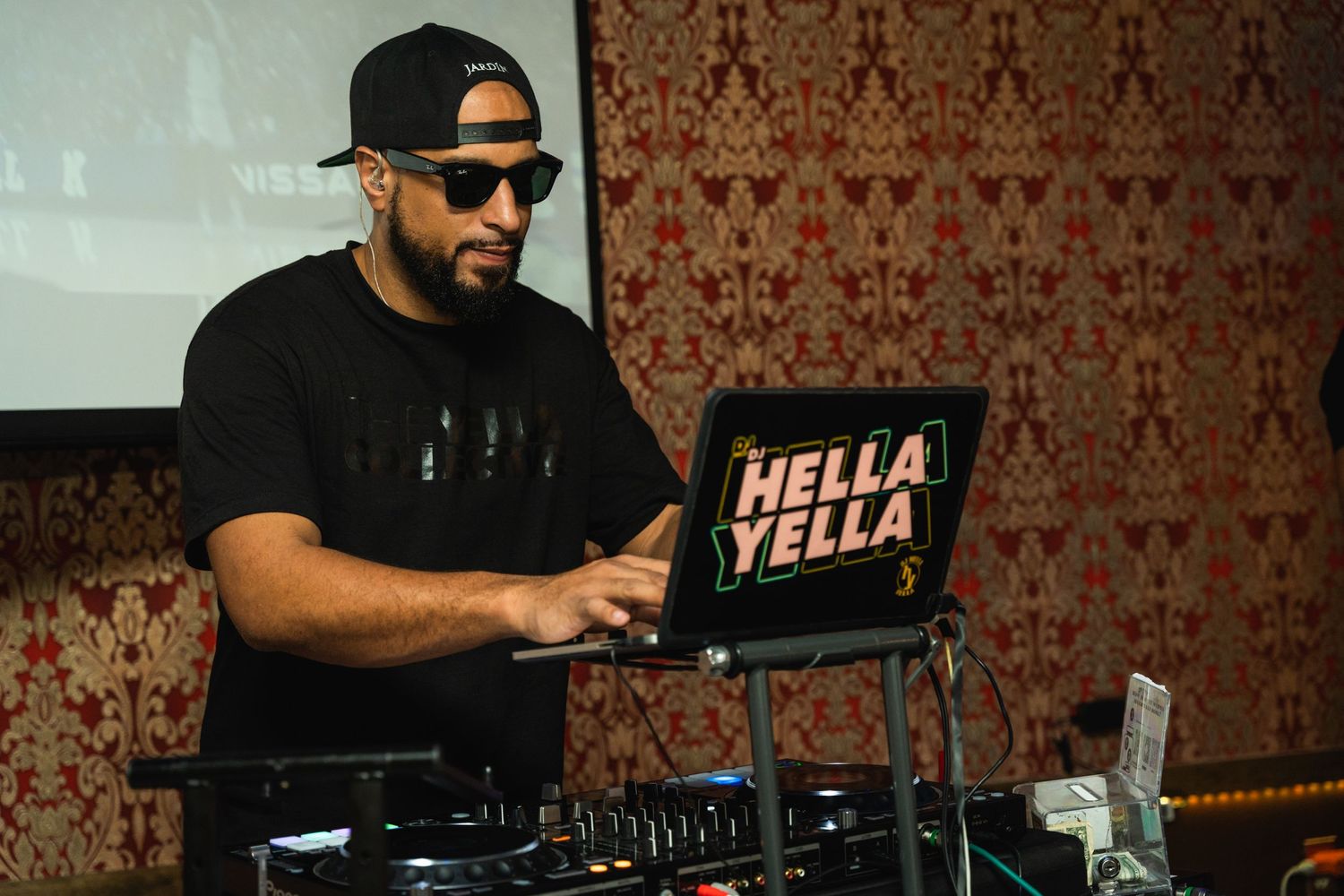 DJ performing with Hella Yella laptop and equipment at event with patterned wallpaper background.