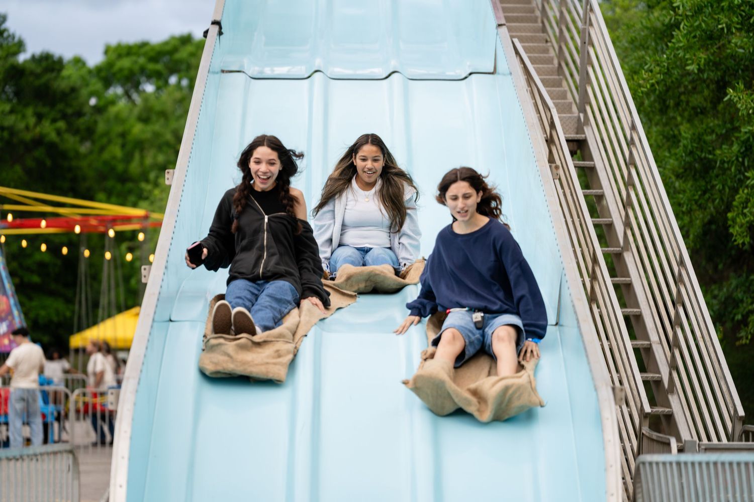 Three girls laughing and sliding down a blue amusement park slide on burlap sacks on a sunny day.