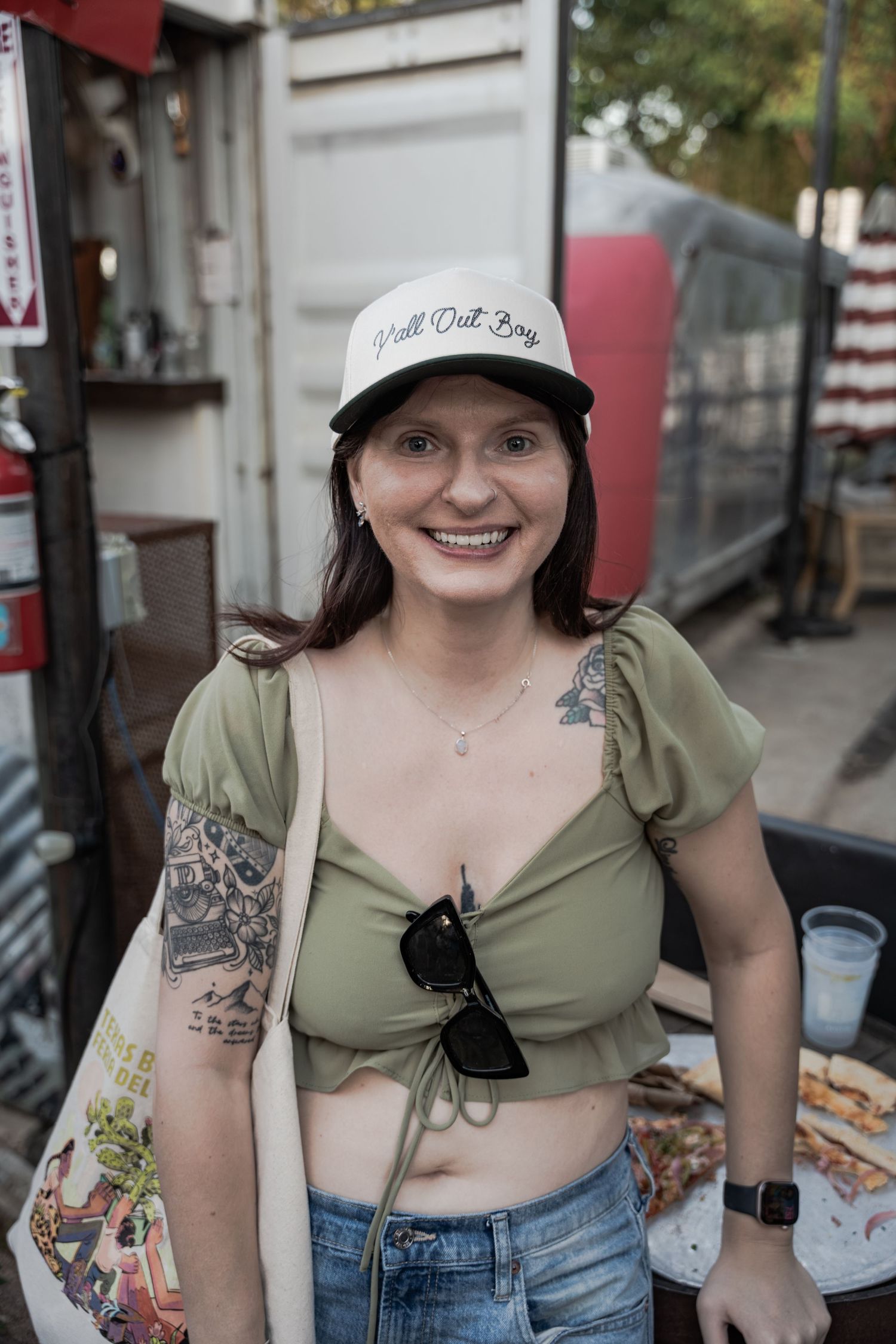 Smiling woman with tattoos wearing a green crop top, white cap, and jeans at an outdoor venue.