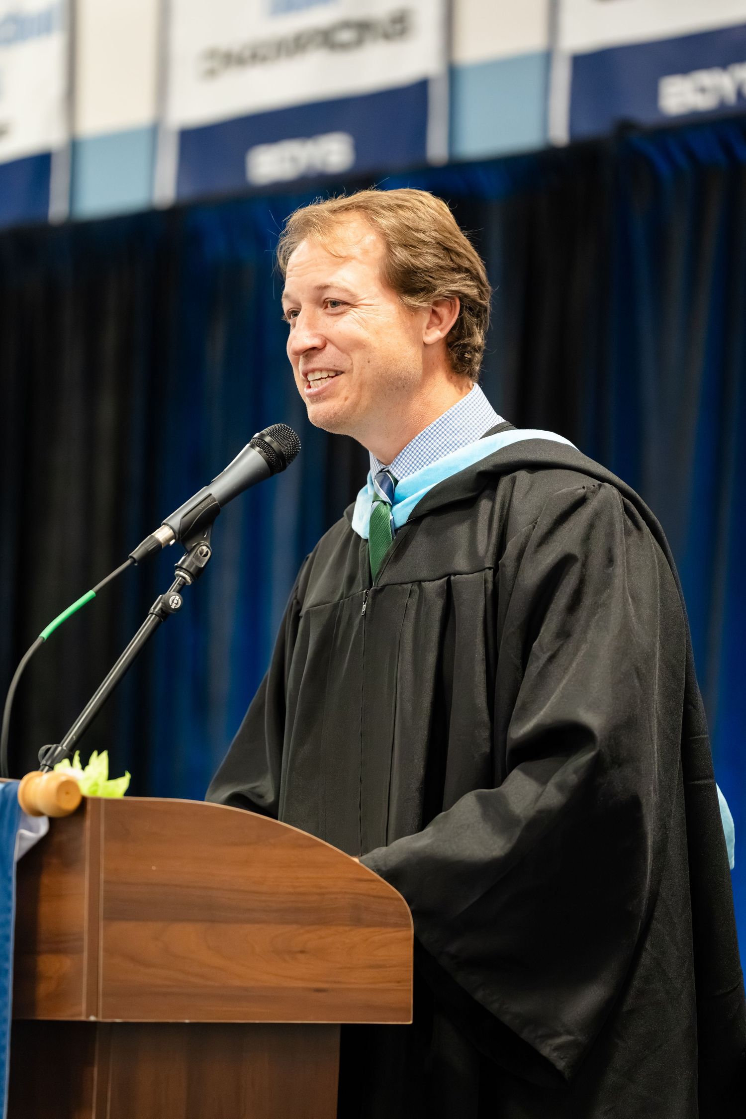 Man in graduation regalia speaking at podium during commencement ceremony in academic setting.