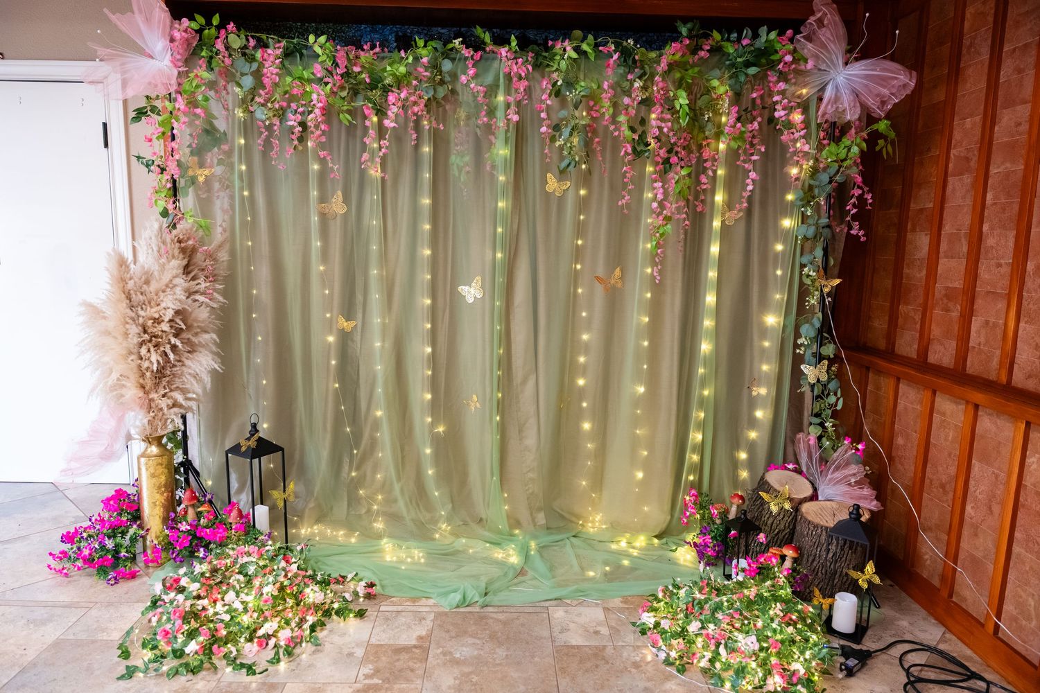 Fairy tale photo booth backdrop with green tulle, string lights, pink flowers, and butterfly decorations for events.