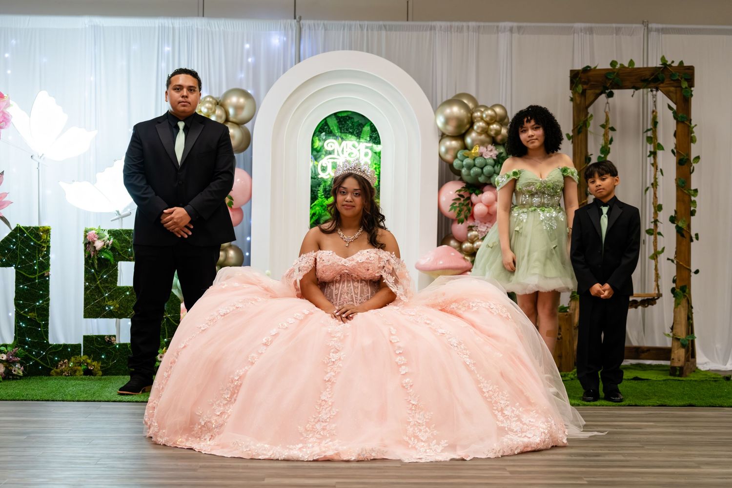 Young woman in pink quinceañera ballgown poses with family at elegant celebration with balloon arch backdrop.