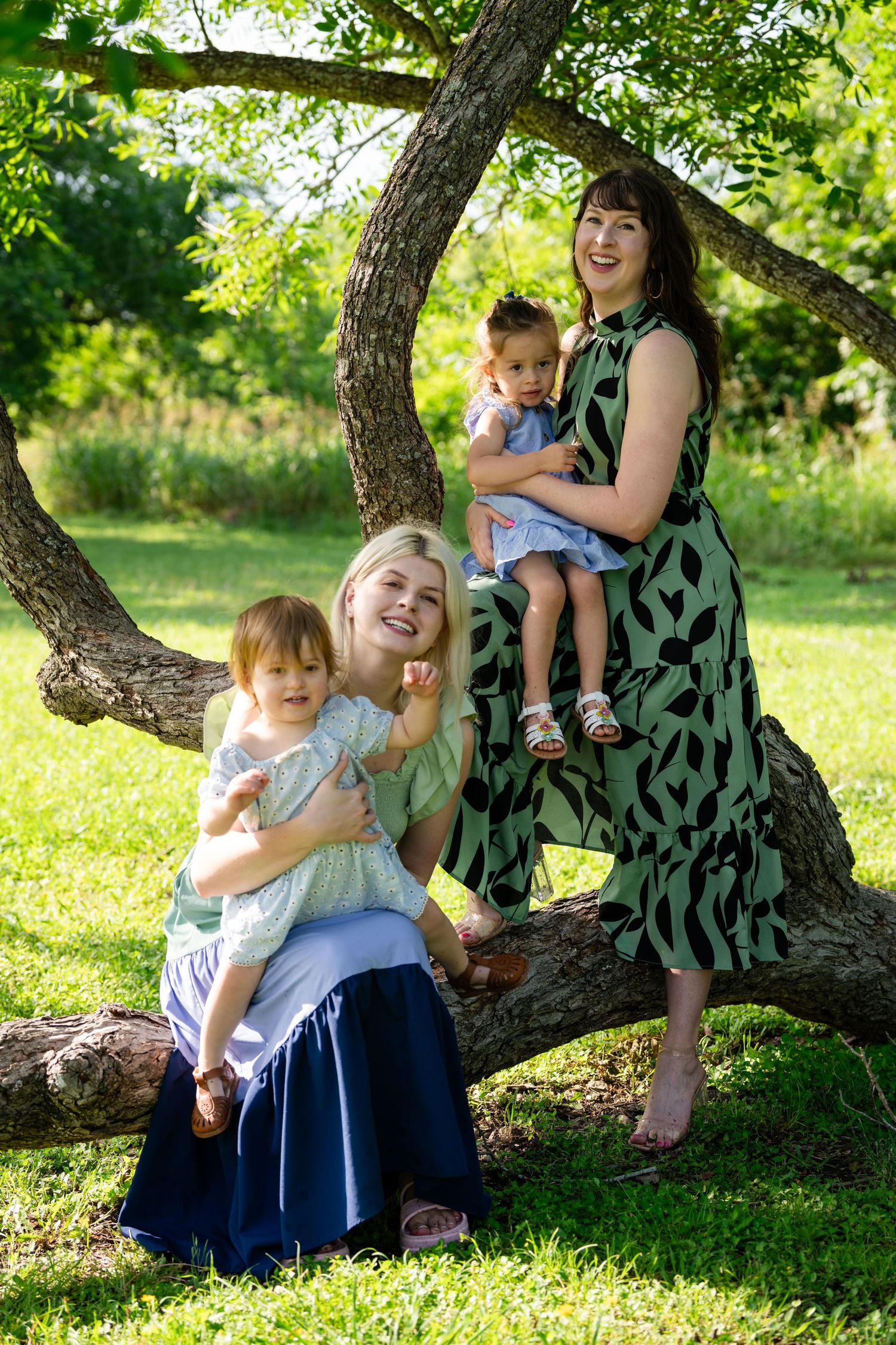 Two women holding toddlers pose outdoors near a large tree on green grass in summer dresses.