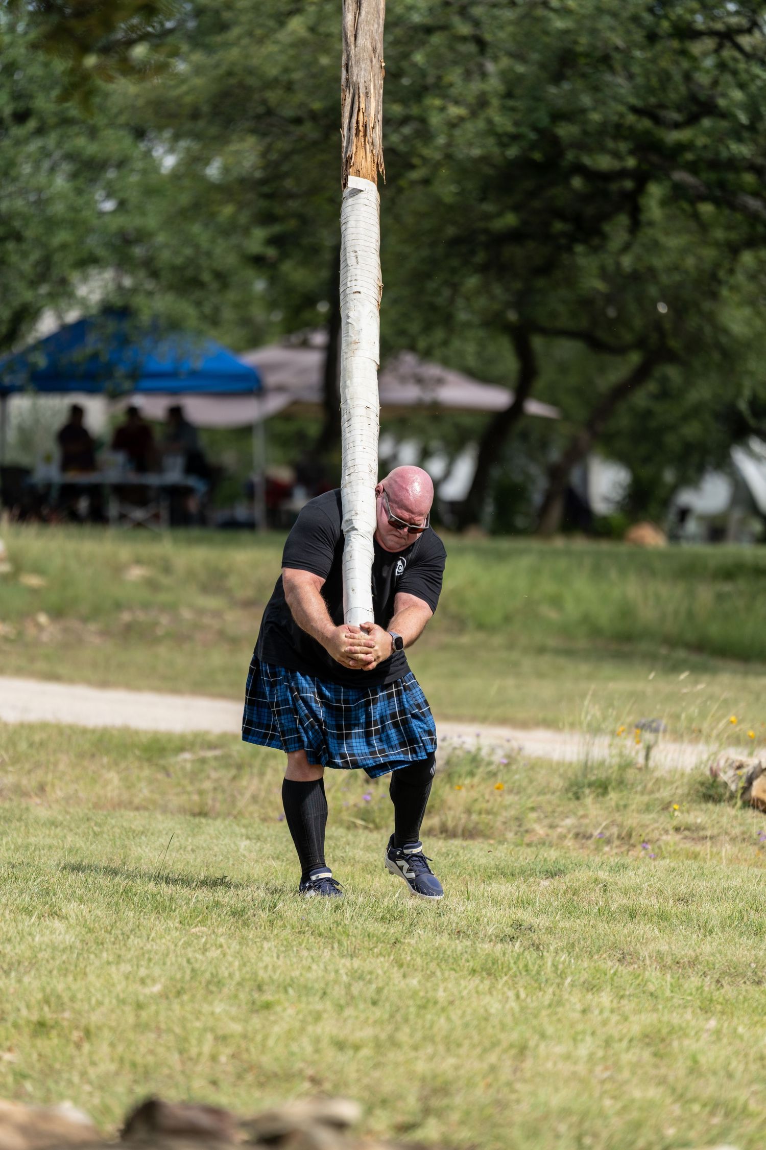 Man in blue tartan kilt tossing a caber at a Scottish Highland Games competition outdoors.