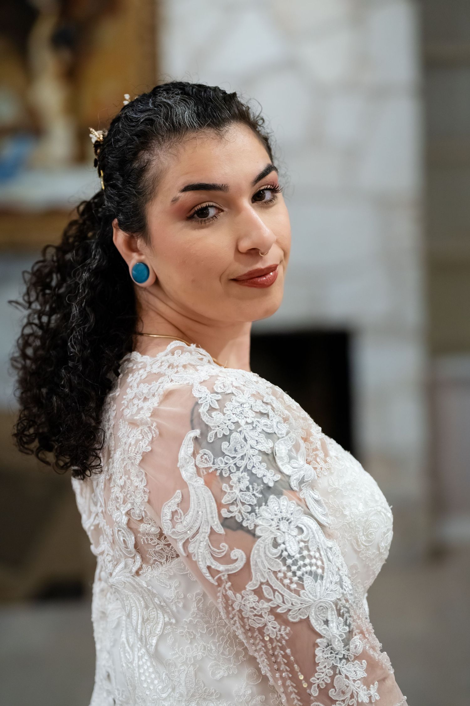 Bride in lace wedding gown with blue earrings and curly hair smiles over shoulder in elegant church setting.