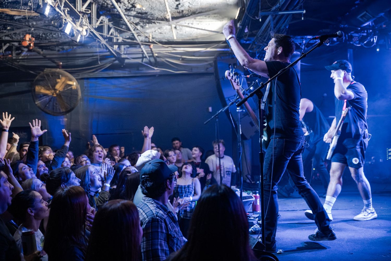 Rock band performing on stage with energetic crowd raising hands at an indoor concert venue with blue lighting.
