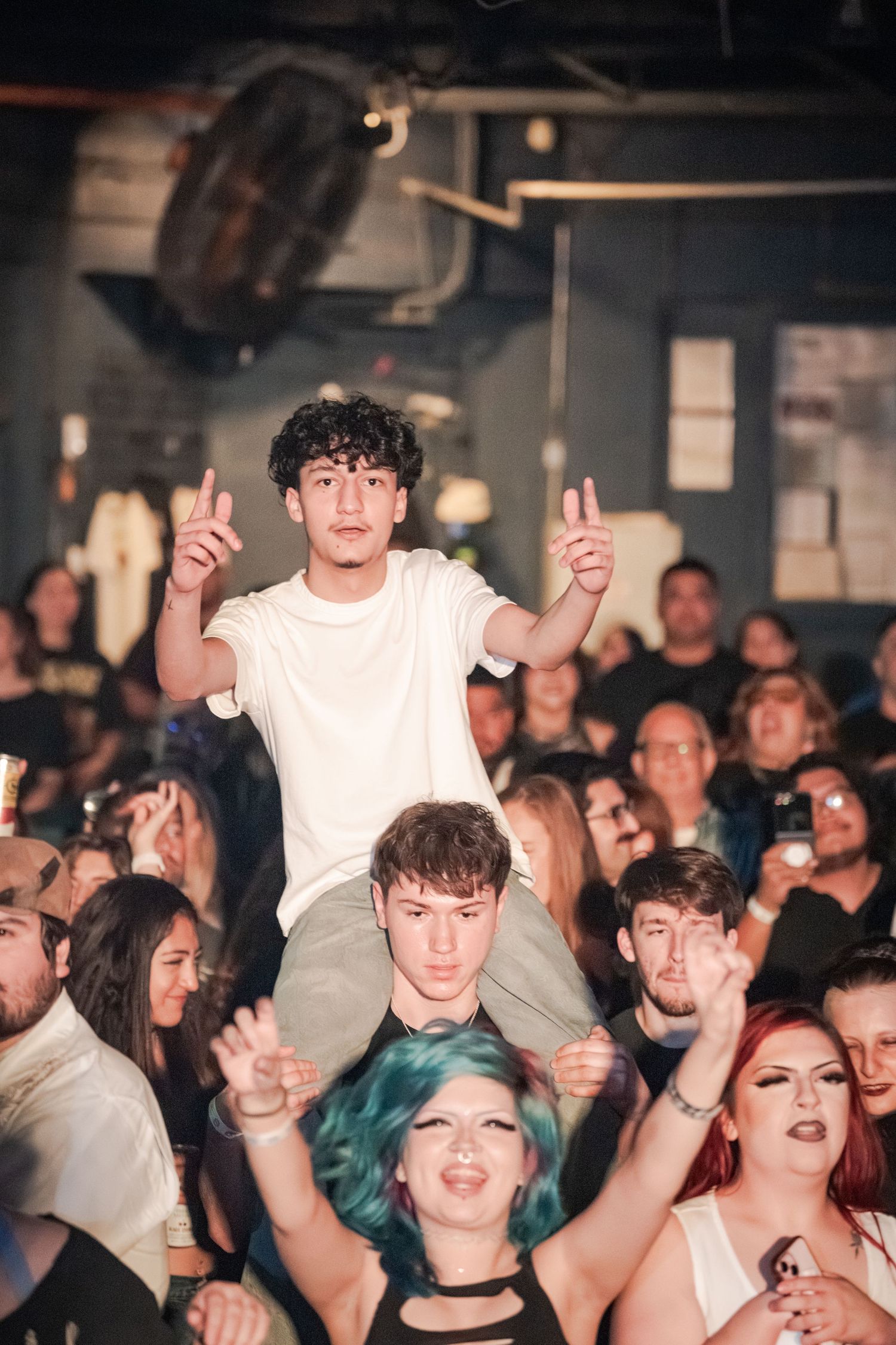 Excited crowd at a concert with a young man in white shirt sitting on shoulders raising fingers.
