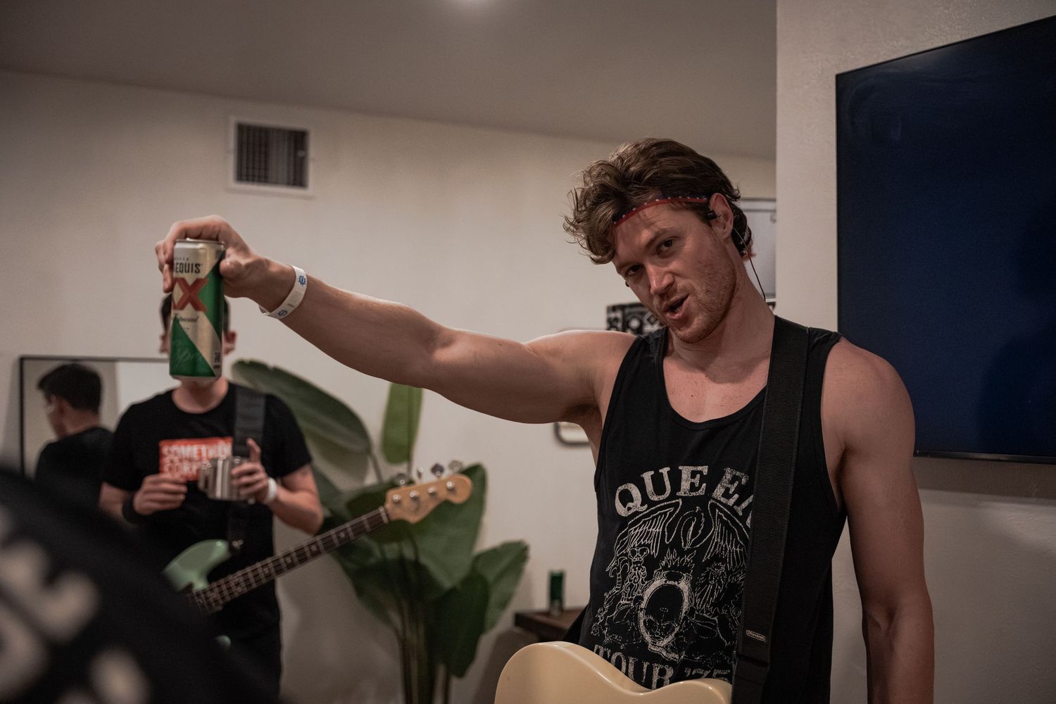 Man in Queen tank top holds up a beer can while another musician plays bass guitar in background.
