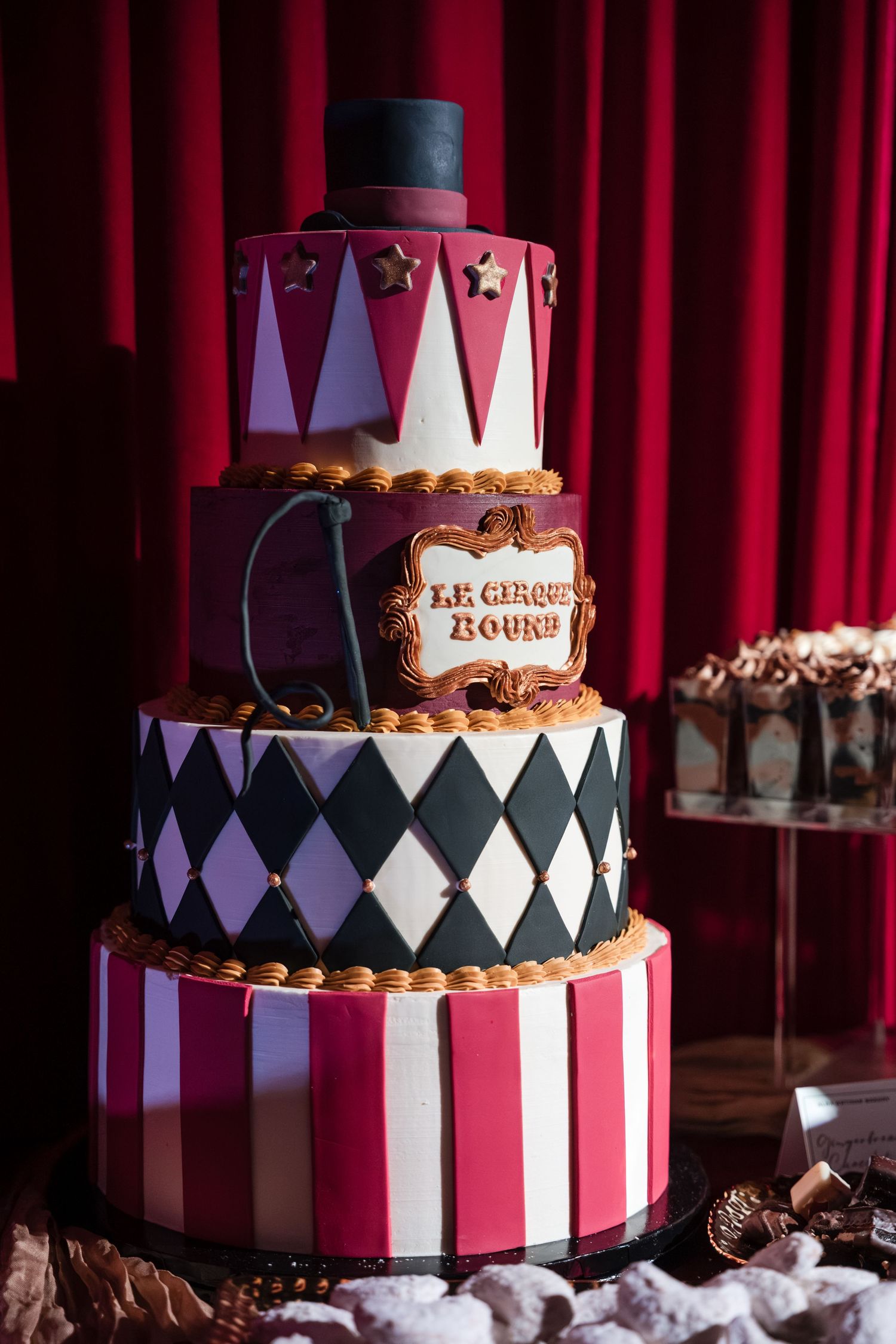 Elegant black, white and red circus-themed tiered cake with diamond patterns and gold crown topper against red curtain backdrop.