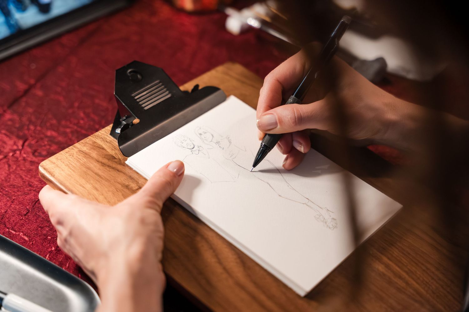 Person writing or drawing on white paper with a black pen on a wooden surface with red tablecloth in background.