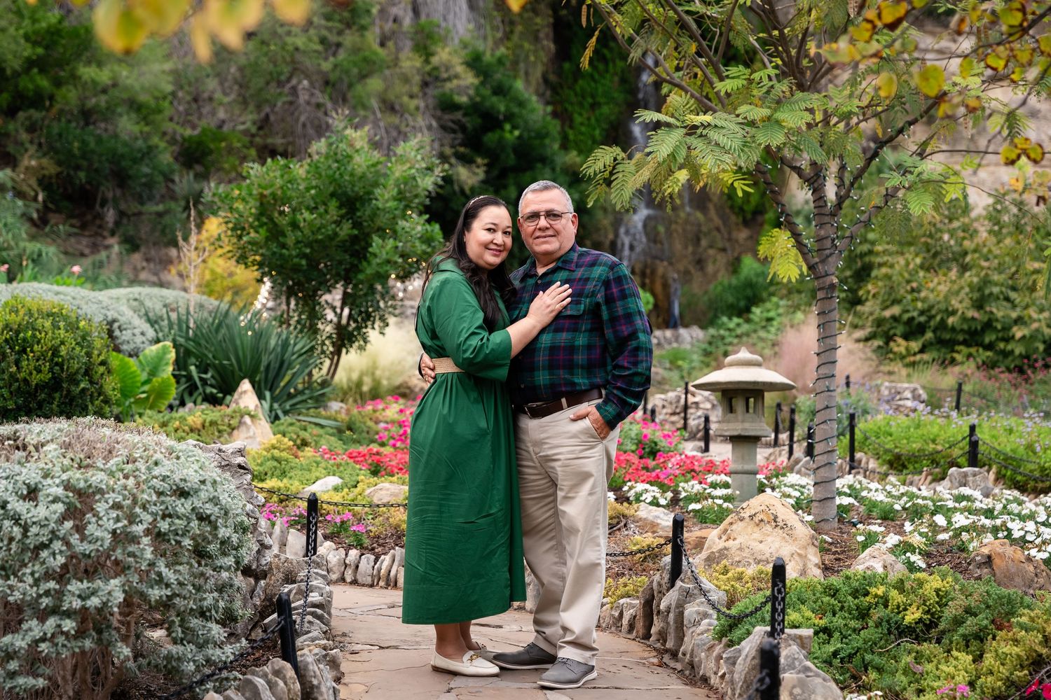 A couple poses together in a lush garden setting wearing coordinated green and gray outfits for outdoor photos.