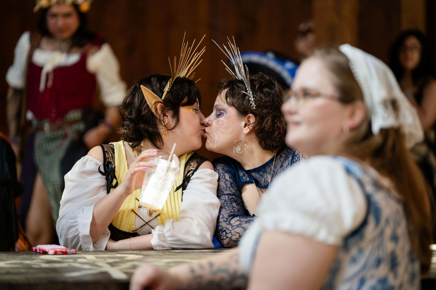 Two people in Renaissance festival costumes share a romantic moment at a wooden table while others look on.