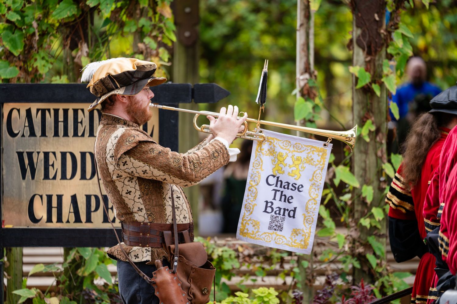 A performer in colonial costume plays a trumpet while holding a Chase the King banner at a chapel outdoor event.
