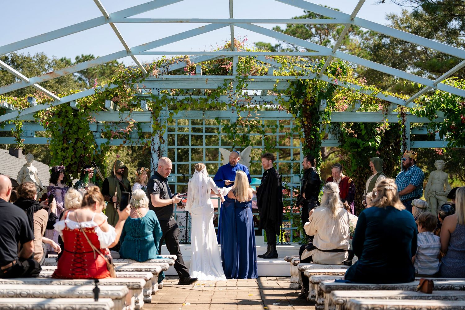 Outdoor wedding ceremony under a glass greenhouse structure with guests seated in white chairs on a sunny day.