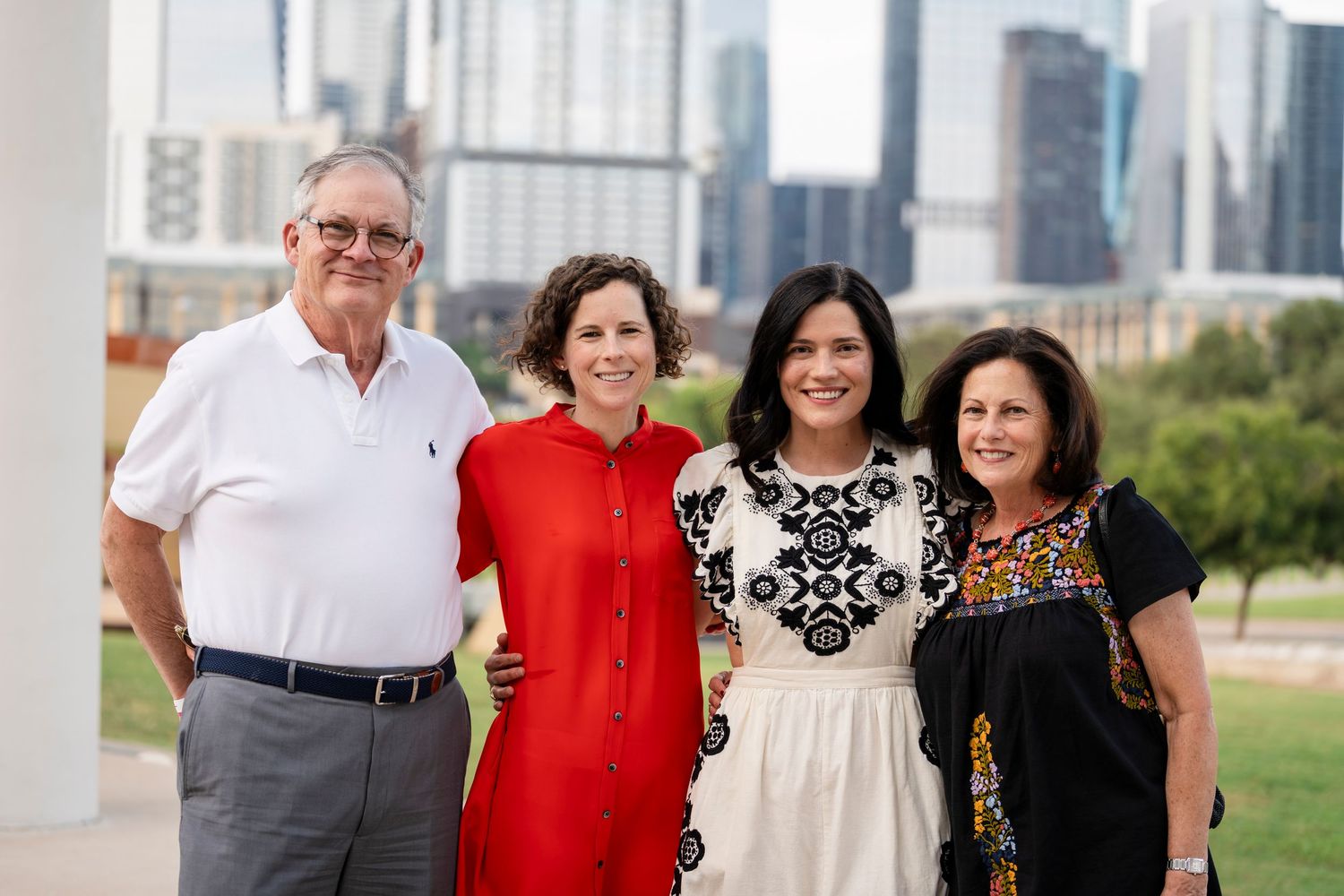 Group of people in casual attire smiling and posing together outdoors with a city skyline visible in background.
