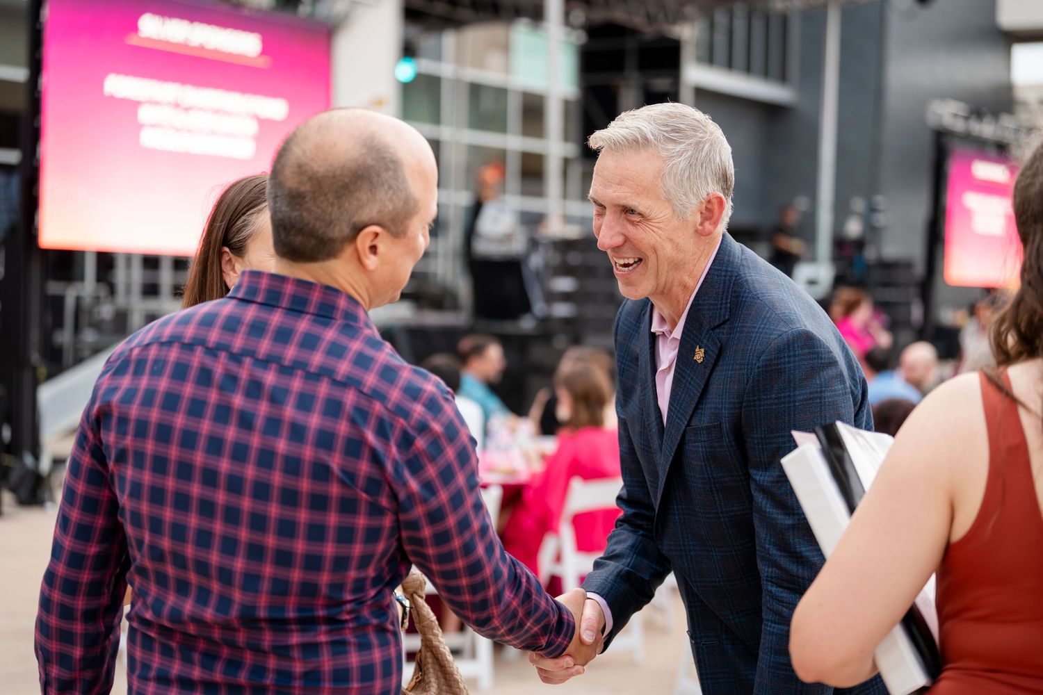 Two people shake hands and smile during a business networking event with pink signage in the background.