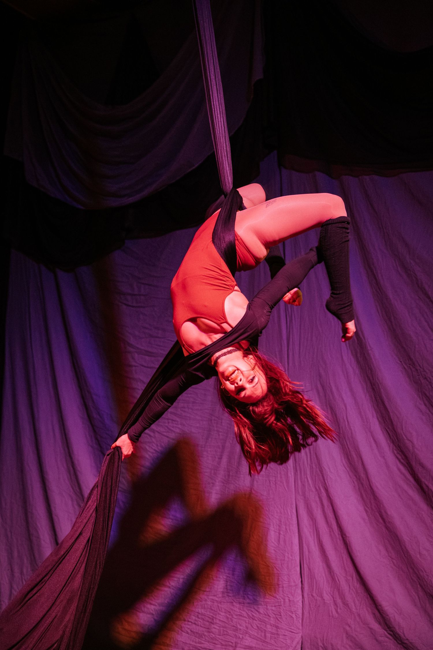 Aerial silk performer in red leotard hanging upside down against purple draped backdrop in dramatic stage lighting.