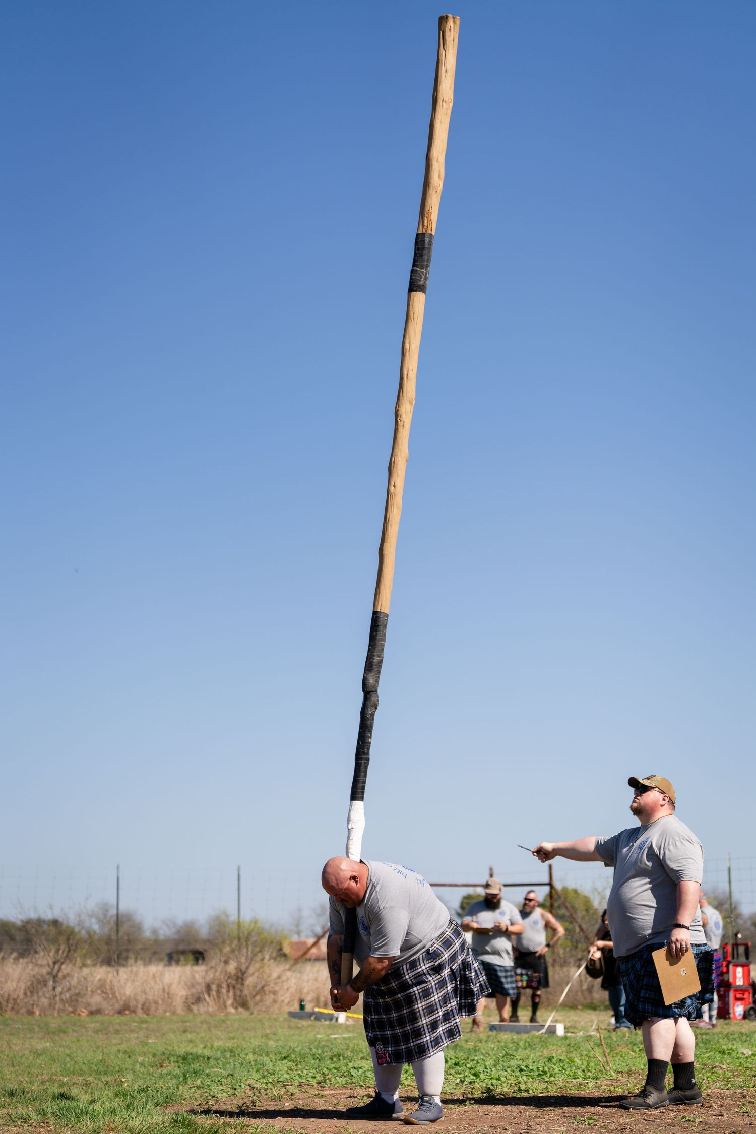 Man in kilt tossing a caber at a Highland Games competition, with a judge observing nearby under a clear blue sky.
