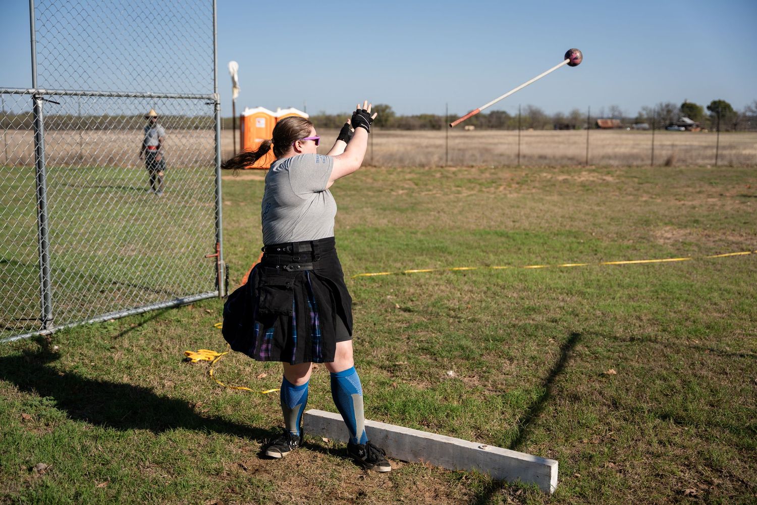 Person in kilt and blue socks throwing hammer at Highland Games competition outdoors.