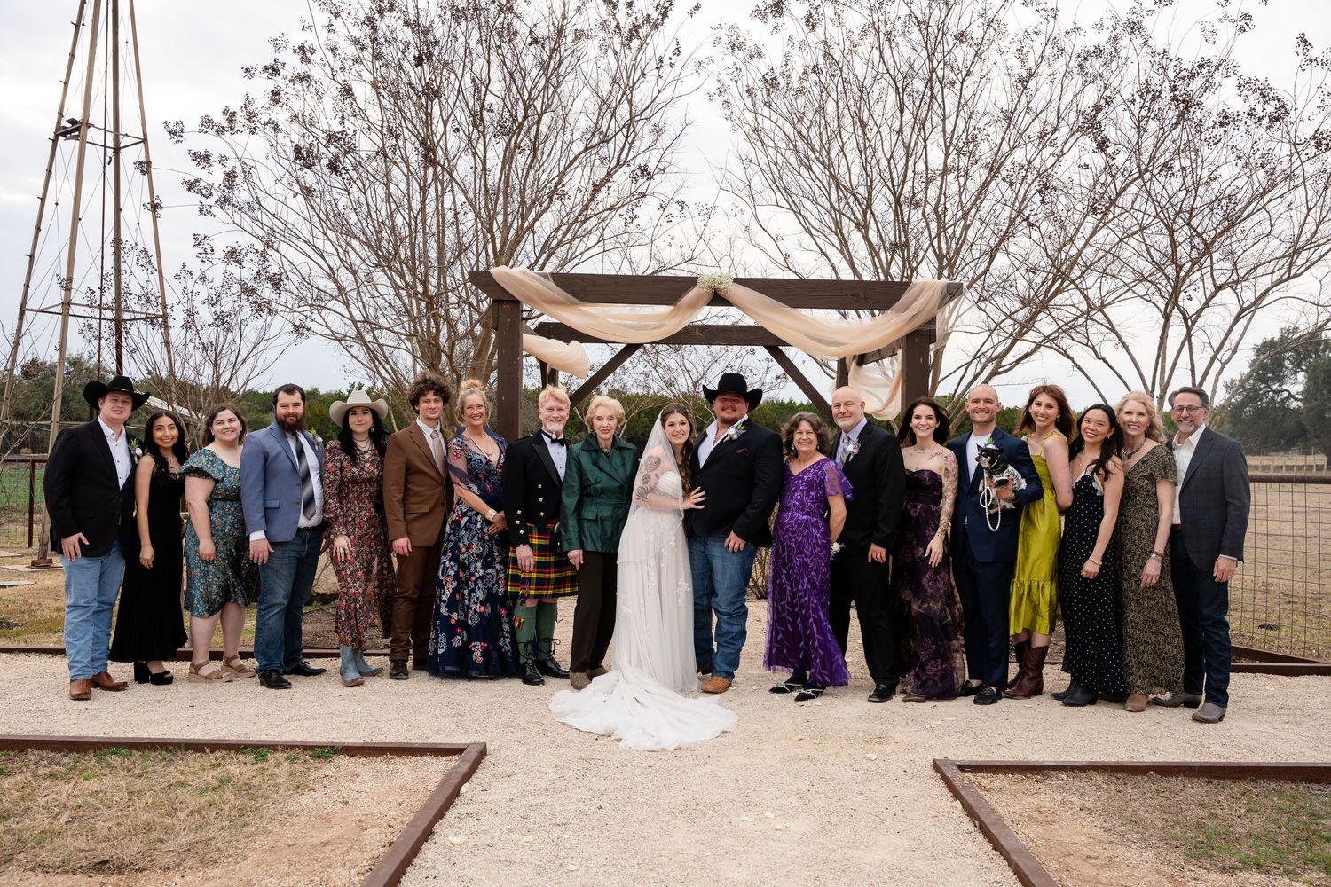 Wedding party poses outdoors under rustic arch with bare trees at country venue.
