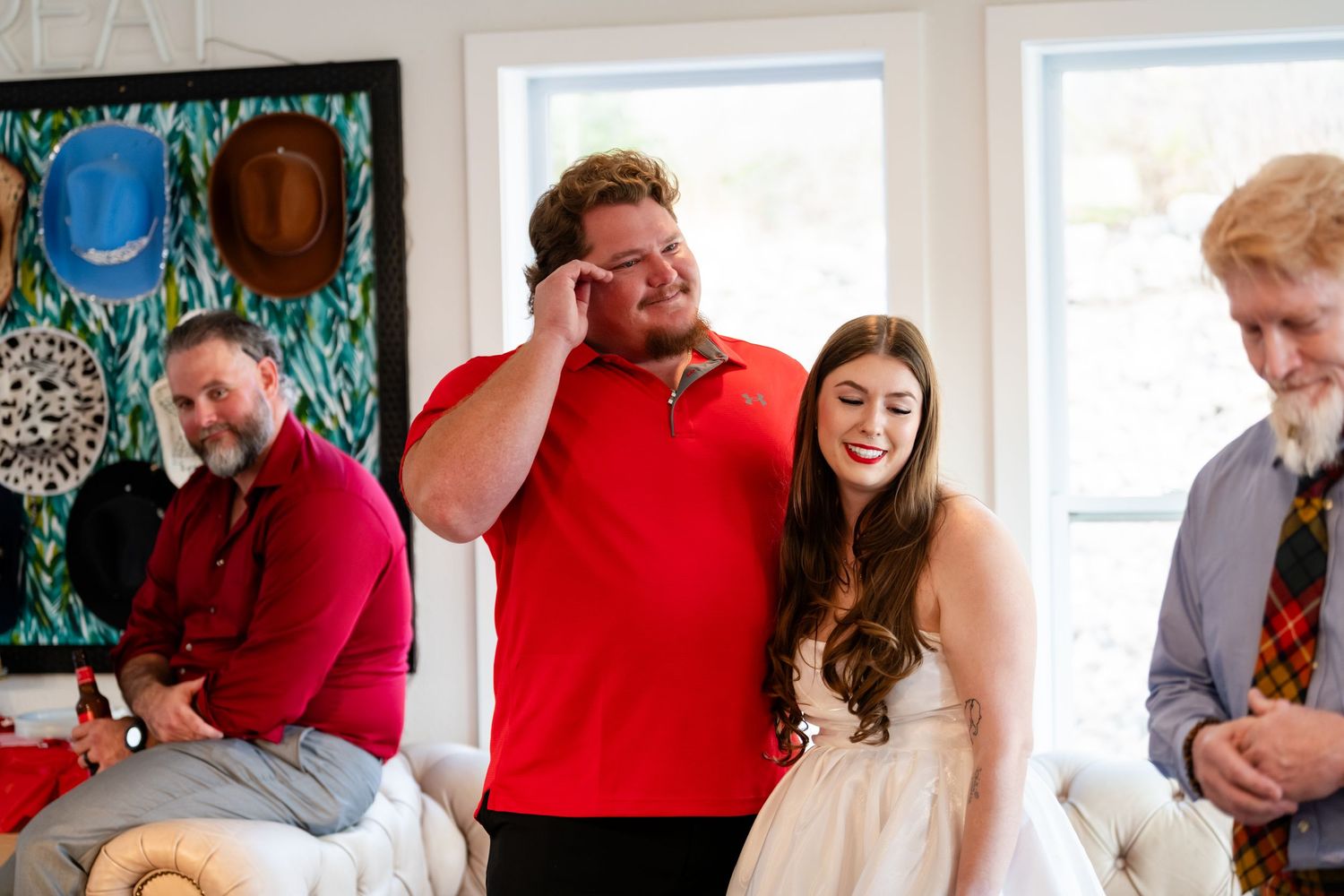 Bride in white dress laughs with guests at casual wedding celebration indoors with colorful artwork on wall.