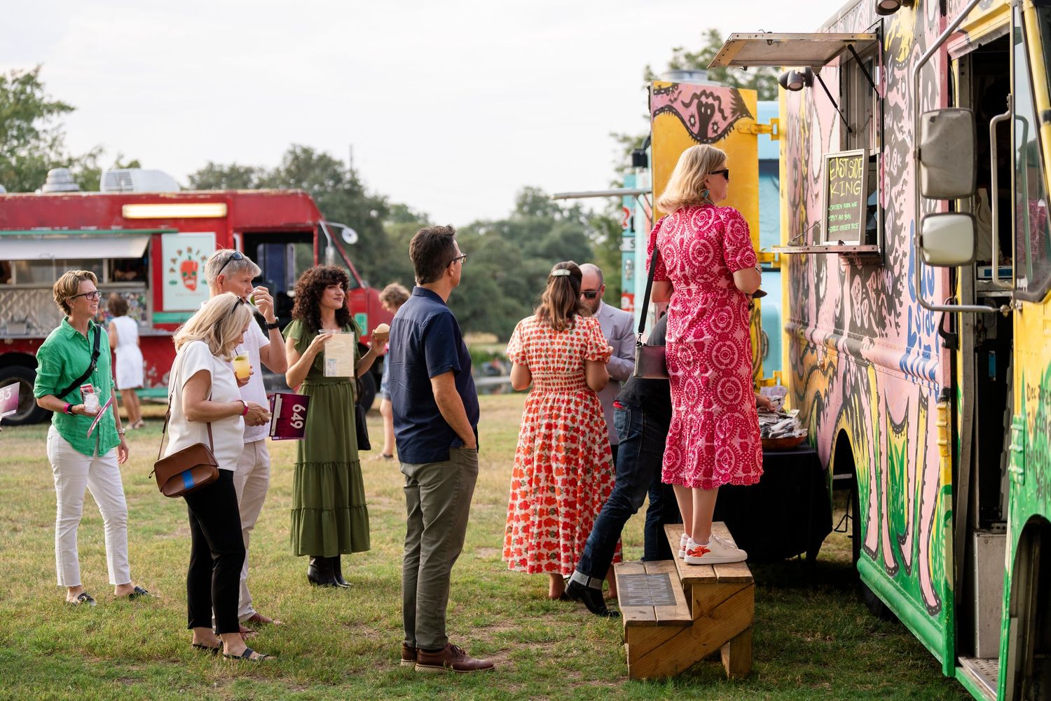 People in colorful dresses and casual attire gather outdoors at a social event near a painted wall.