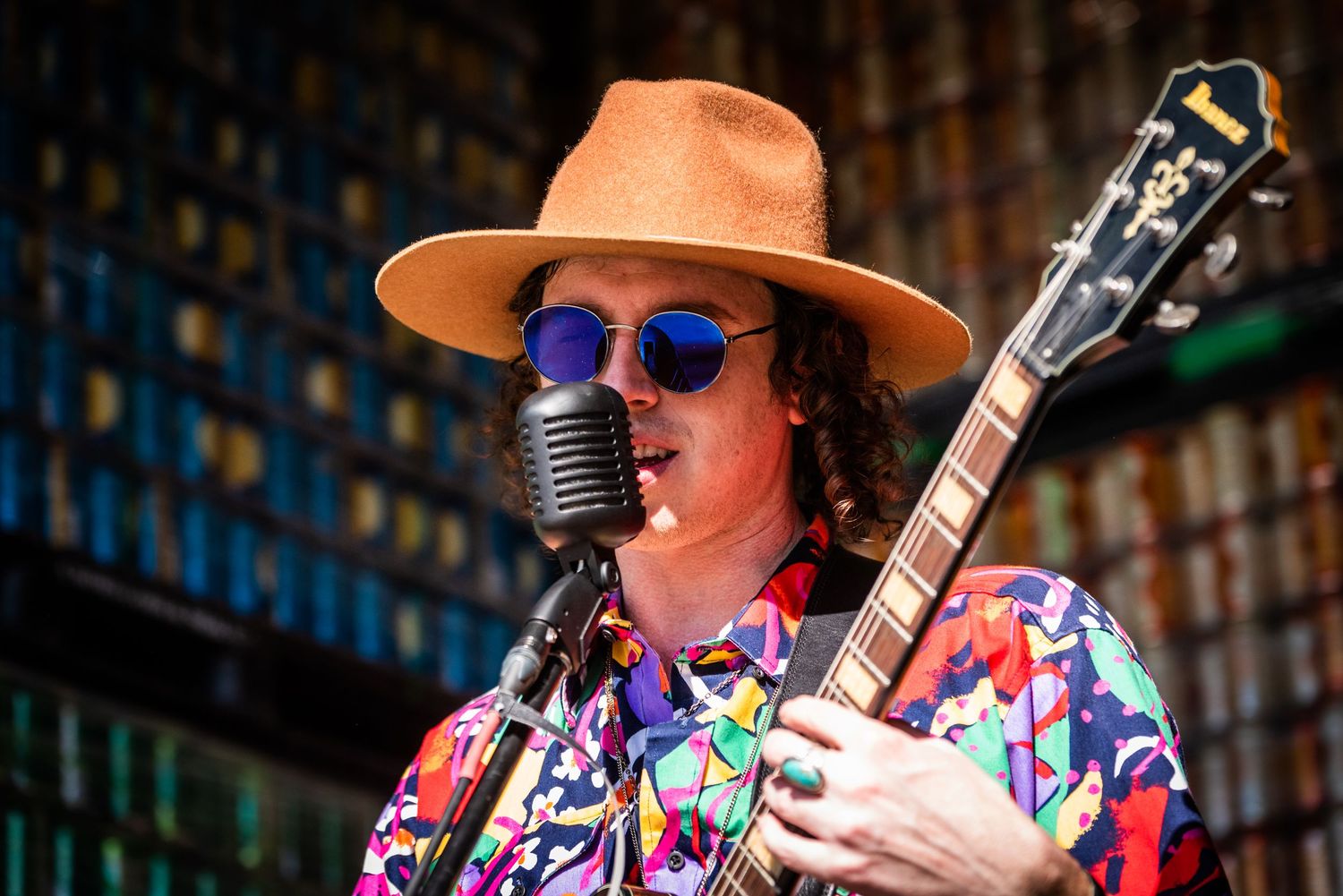 A musician in a colorful shirt and brown hat performs with an electric guitar on stage at a live concert venue.