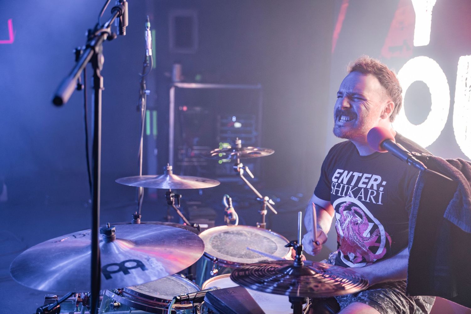 A drummer performs energetically on stage with a drum kit, illuminated by blue and red stage lighting.