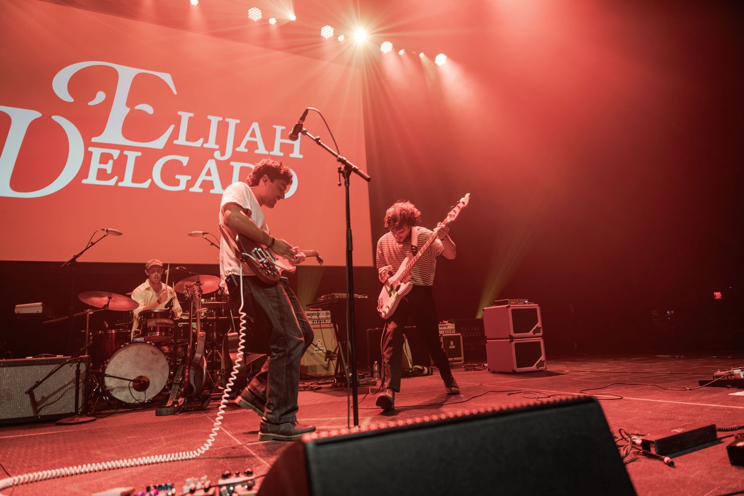 Elijah Delgado band performing live on stage with guitars and drums under red concert lighting.