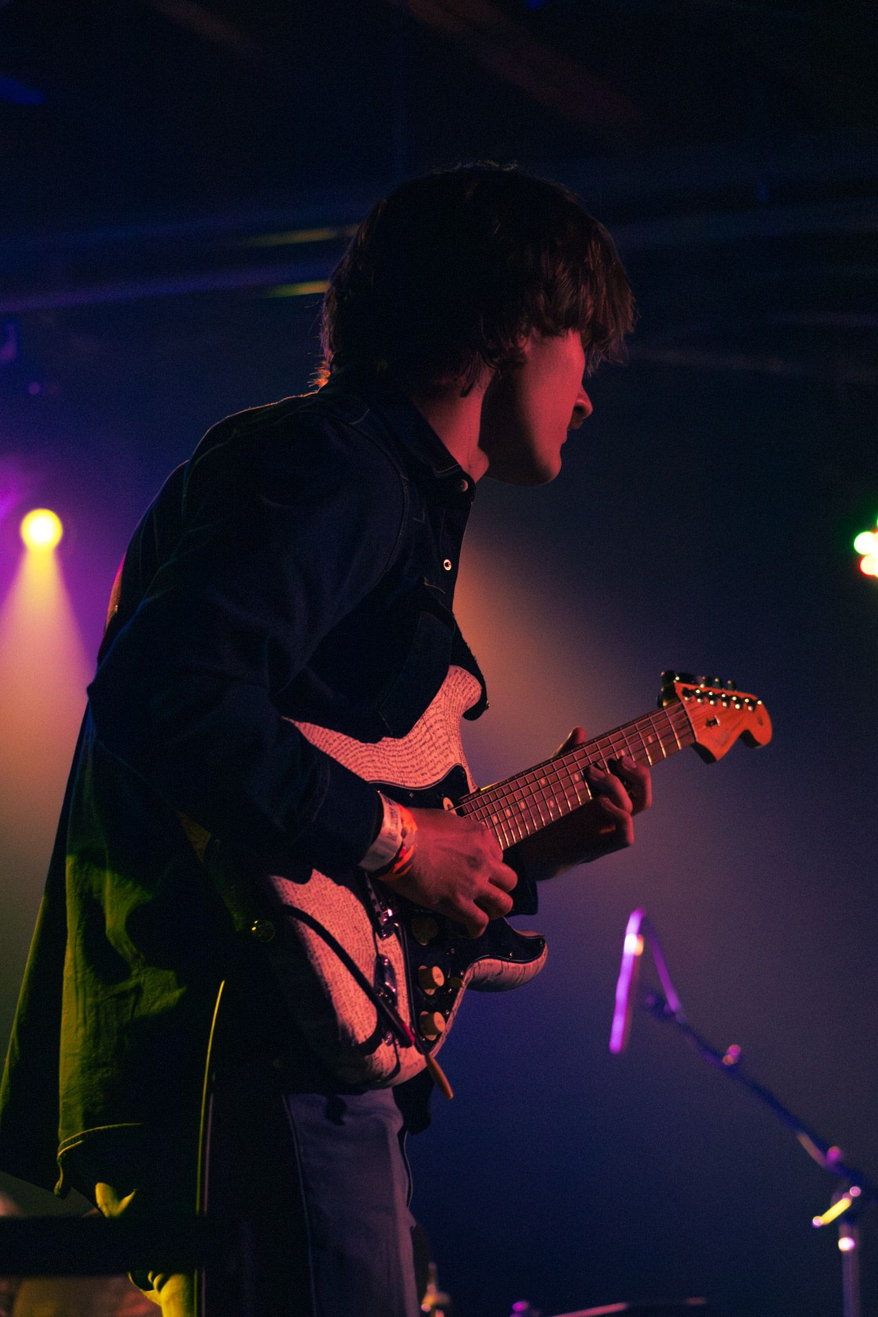 Male guitarist performing on stage under colorful purple and yellow stage lights in a dark venue.