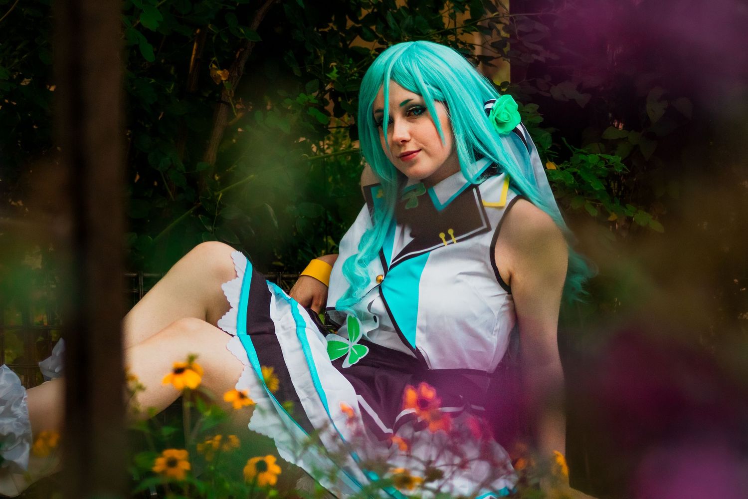 A cosplay figure in turquoise wig and white dress sits among flowers in a dreamy forest setting.