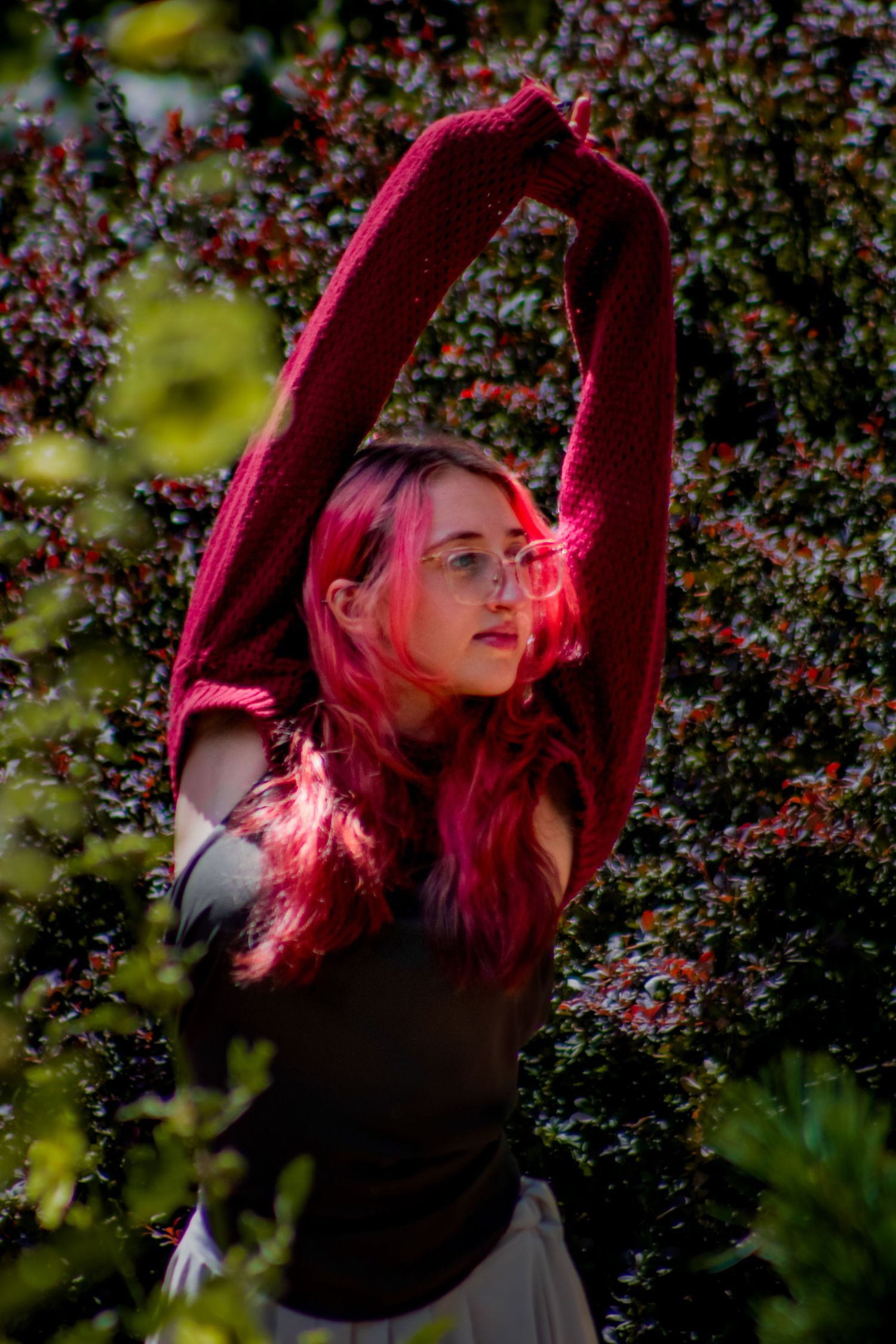 Figure with bright hair stretches in a red sweater among flowering bushes.