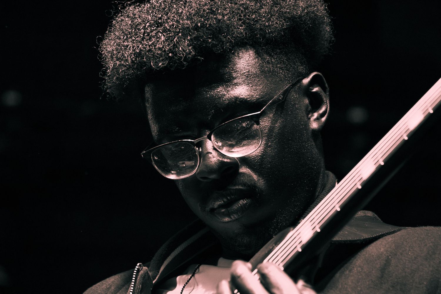 A focused Black musician with an afro and glasses plays electric guitar in a moody, dark-toned performance shot.