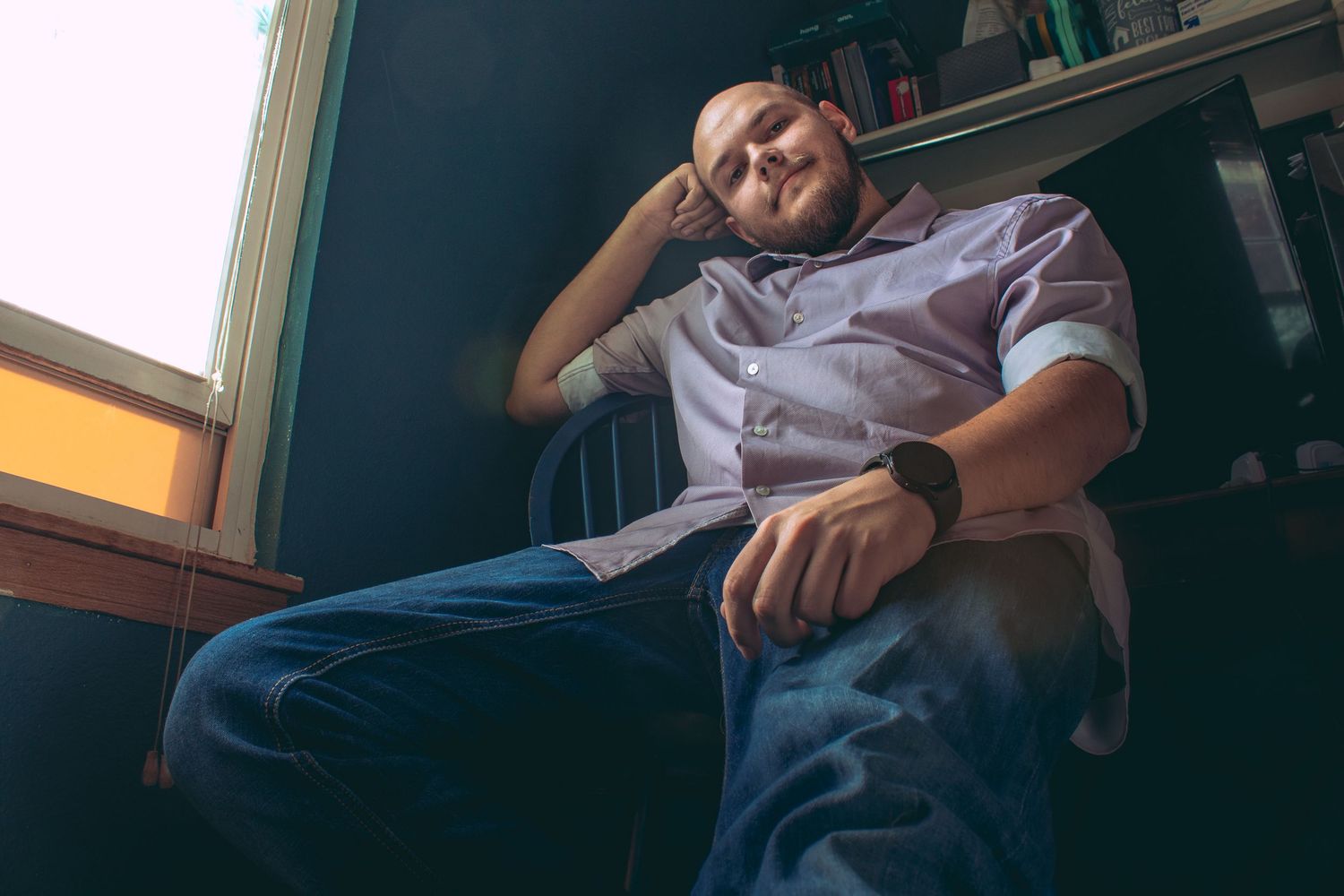 Figure rests thoughtfully in chair near window with dark teal walls and bookshelf.