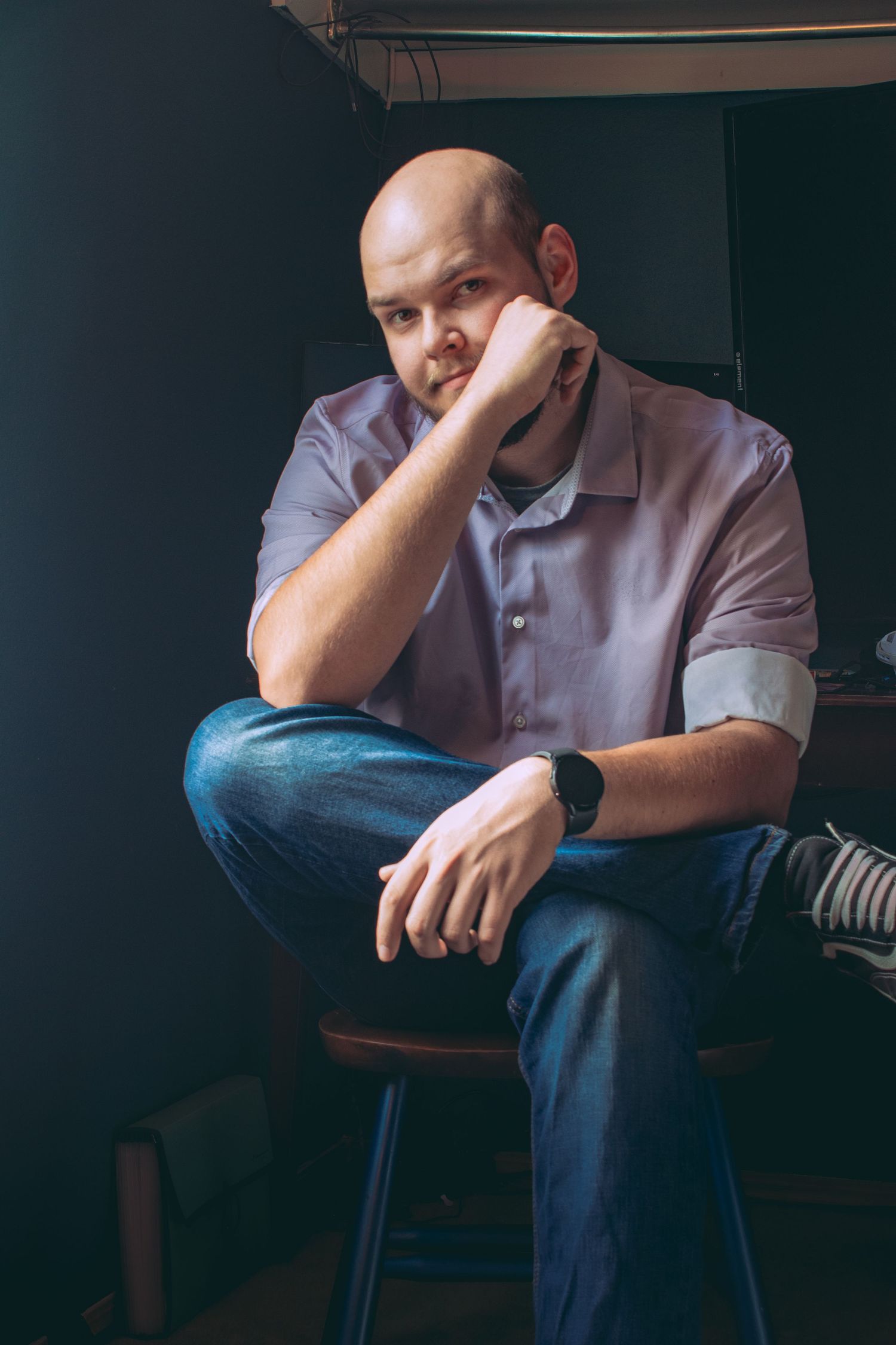 Person sits pensively in wooden chair against dark walls in moody lighting.