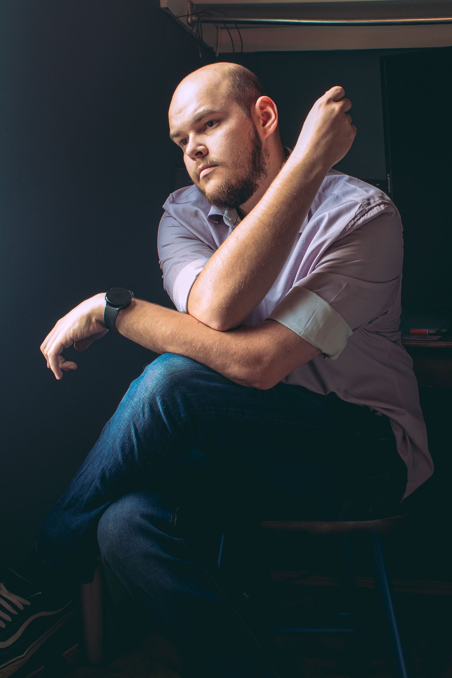 Person in casual attire sits contemplatively in wooden chair against dark walls.