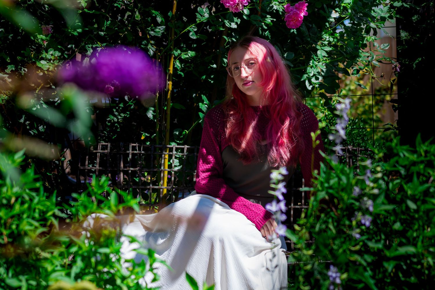 Person wearing red sits amidst lush greenery and purple blooms in a sunlit garden.