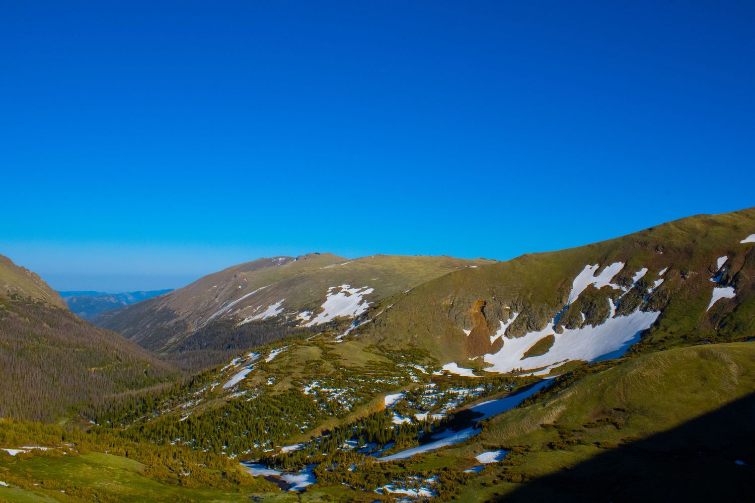 Alpine landscape with patches of snow and small mountain lakes dotting the rolling terrain under bright blue skies.