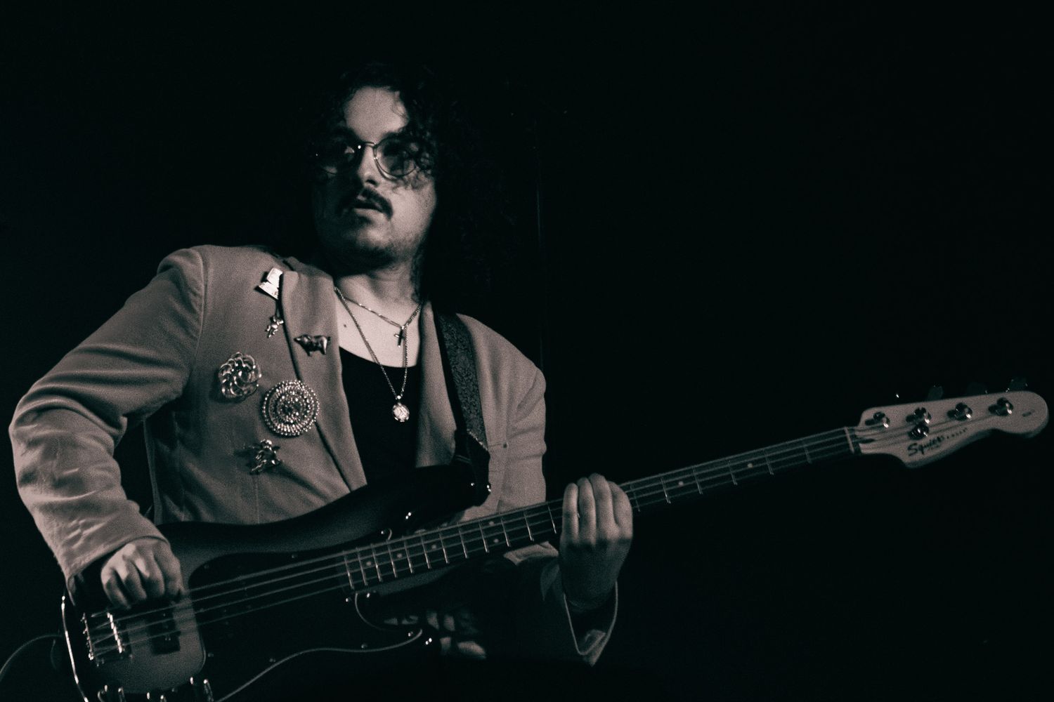 Musician playing bass guitar on stage in black and white, wearing glasses and a jacket with pins.