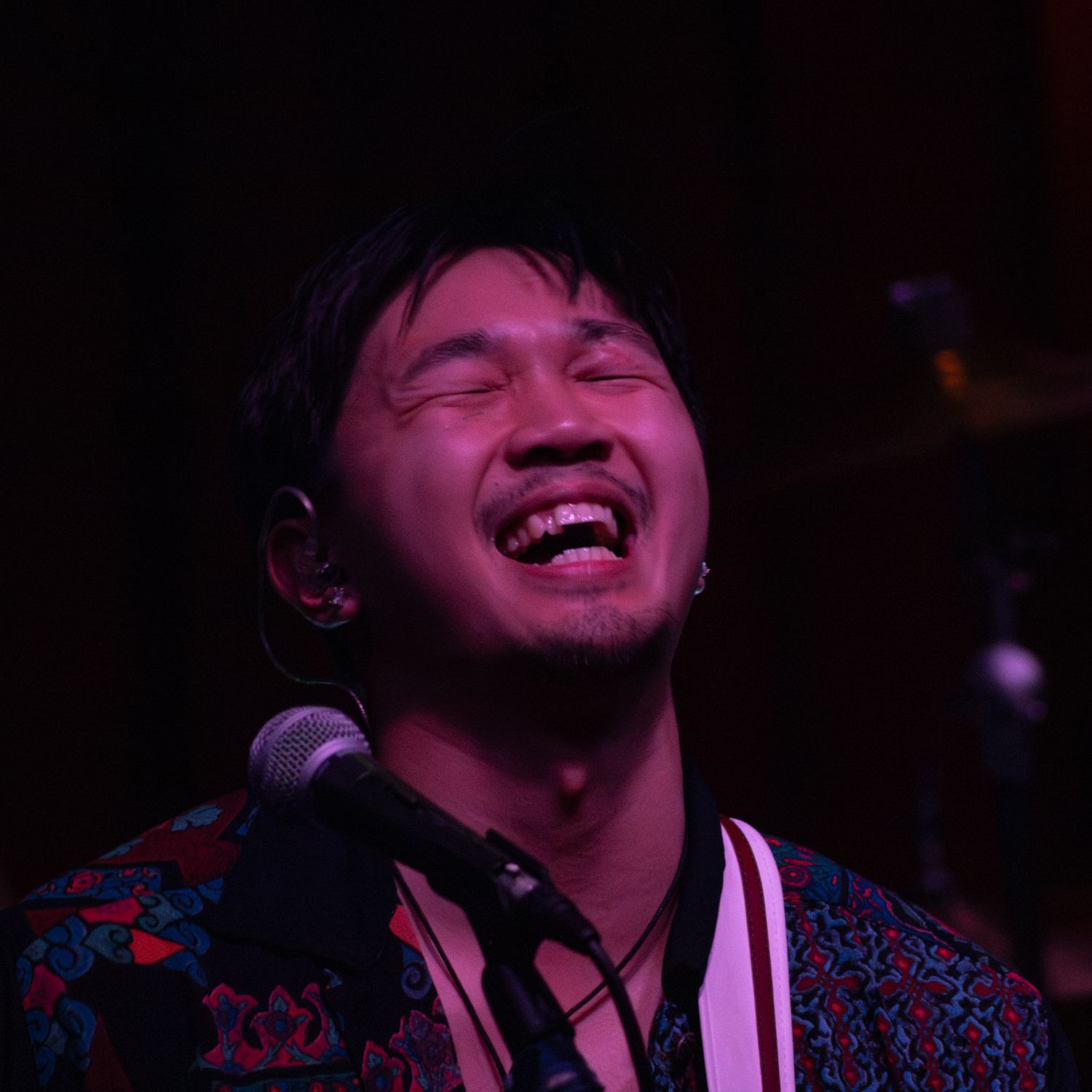 A joyful Asian male singer laughs while performing at a microphone under colorful stage lighting.