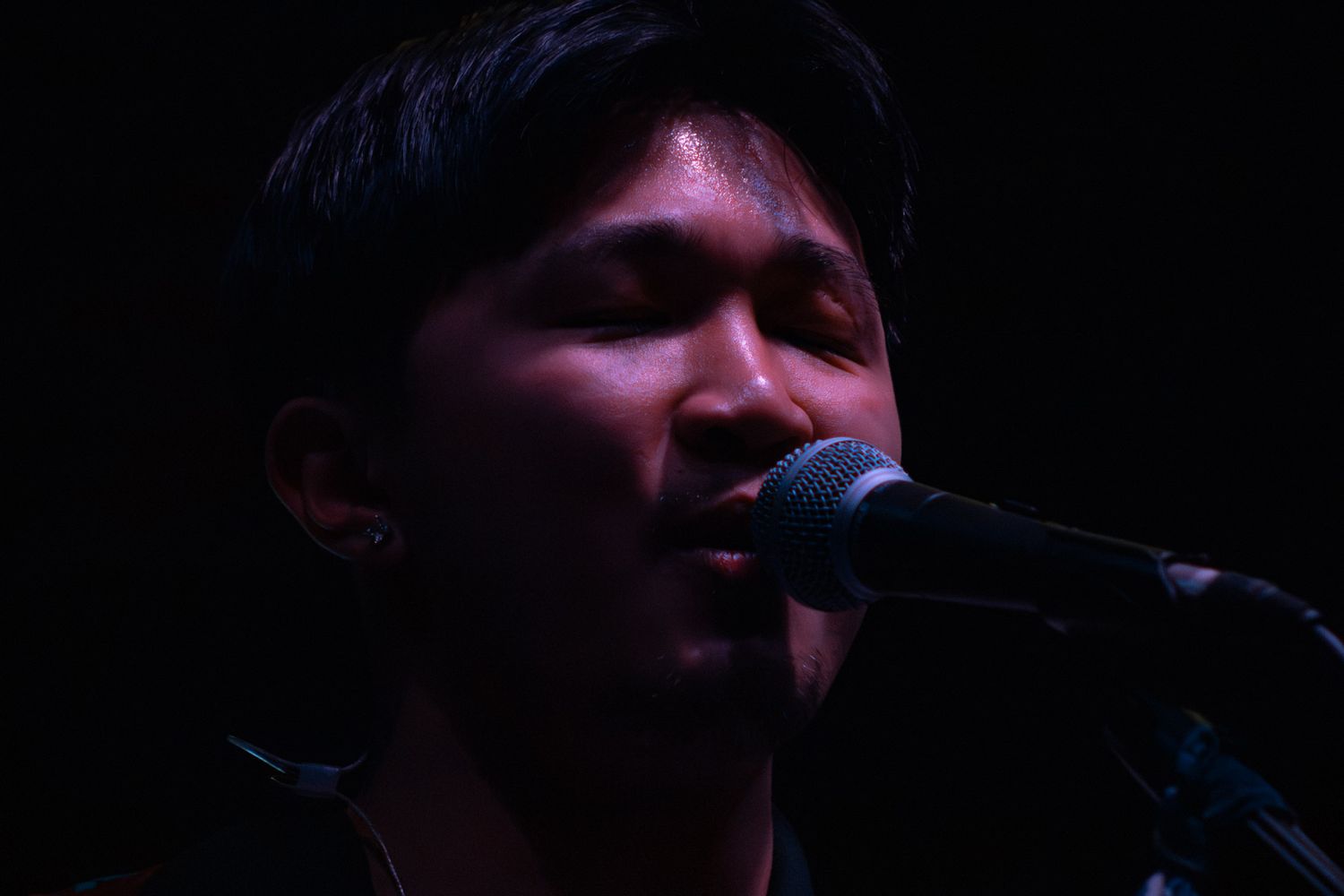 Young male musician performing on stage, singing into a microphone with eyes closed under dramatic dark lighting.