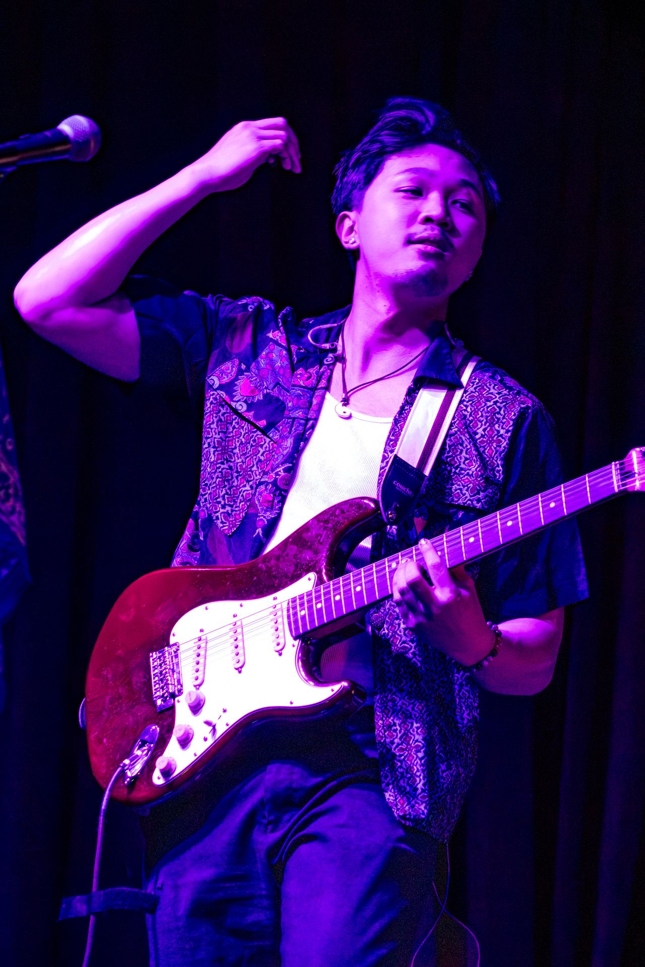 Young Asian guitarist performs on stage with a red Fender Stratocaster under purple lights at a live music event.