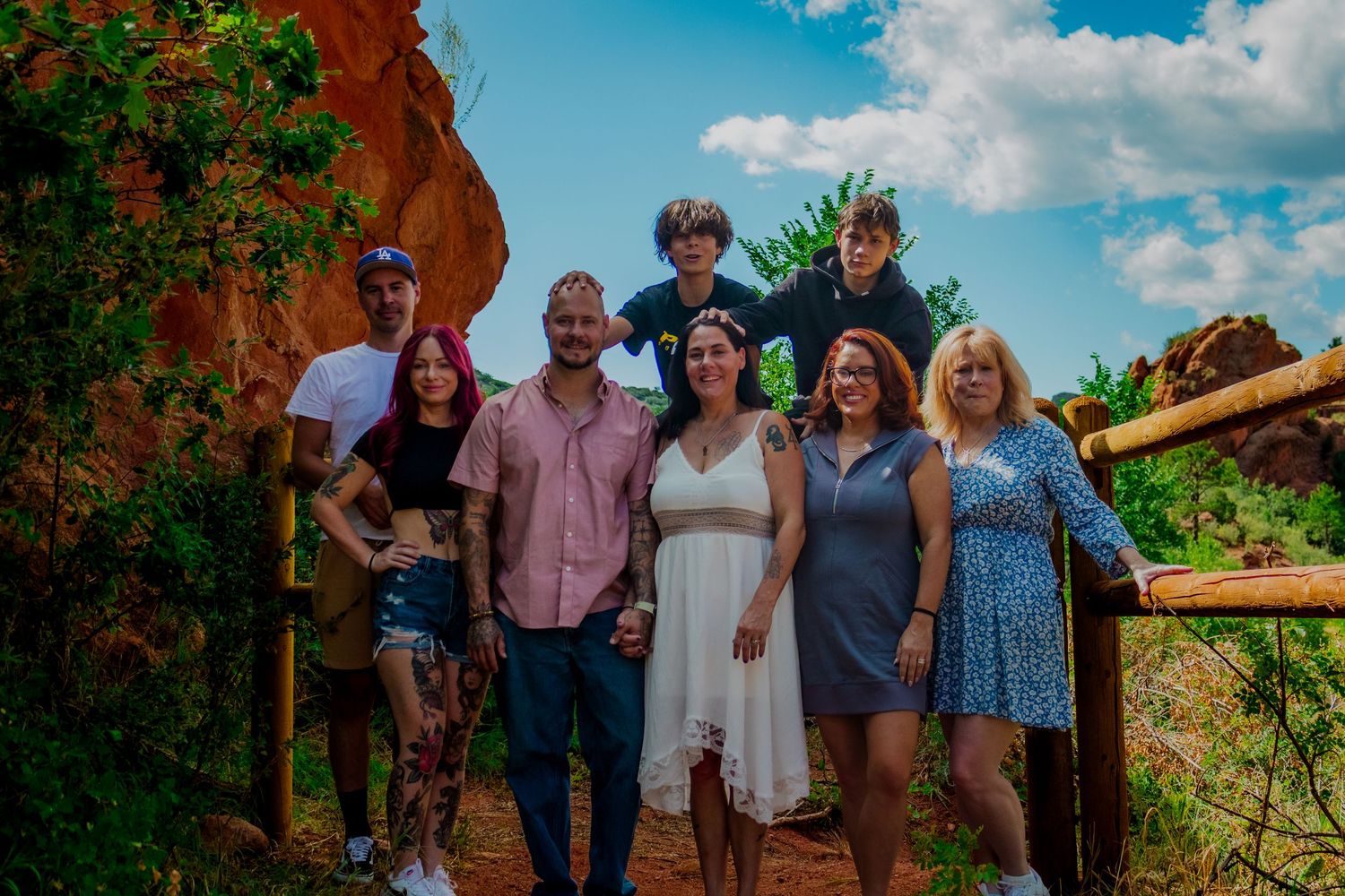 Tourists gathered for a photo along a wooden railing with towering red sandstone cliffs and blue sky in background.