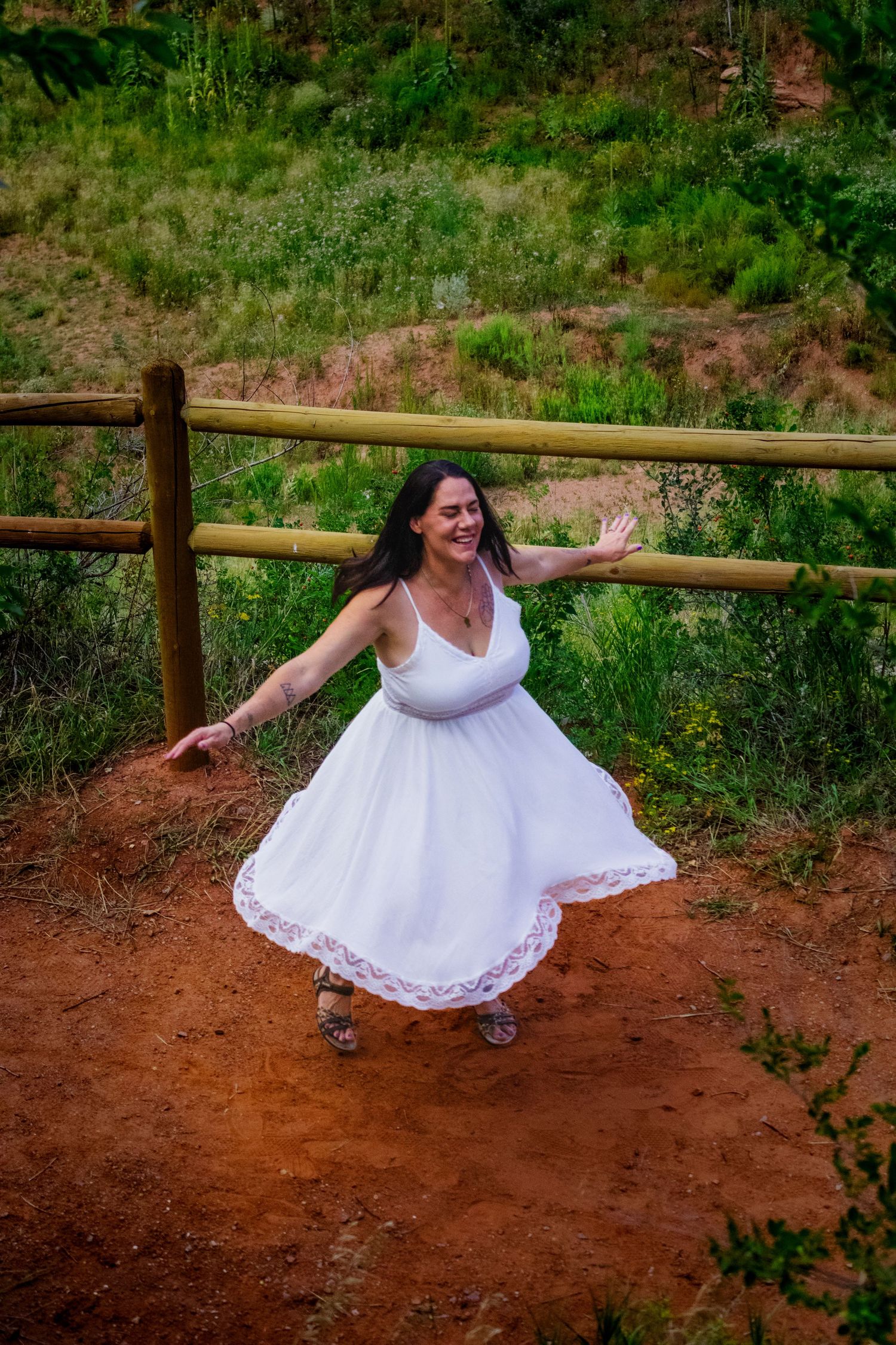 Someone in a flowing white dress spins joyfully on a dirt path with a wooden fence and green hillside in the background.