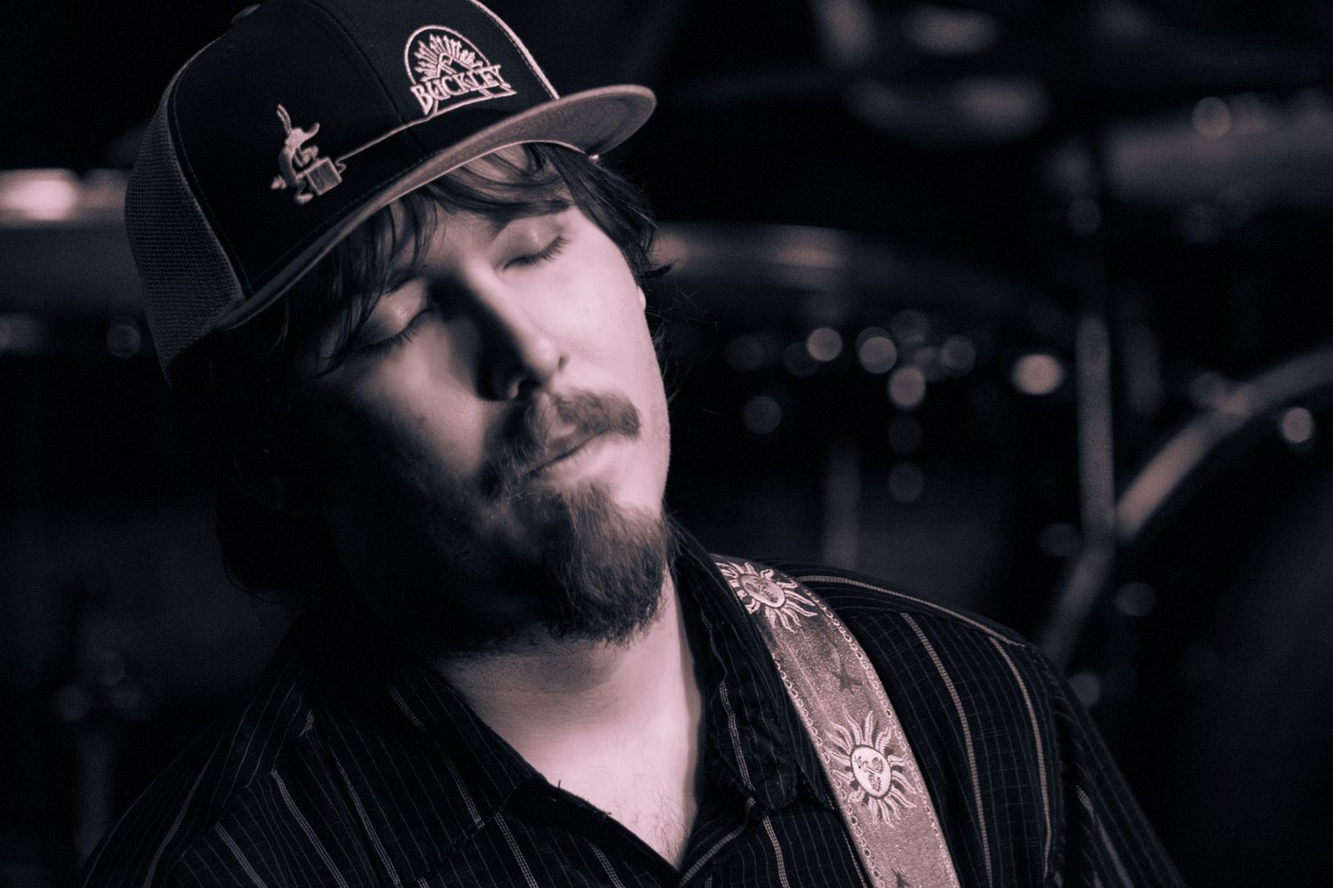 A bearded musician in a snapback hat plays guitar with eyes closed on a dark stage.